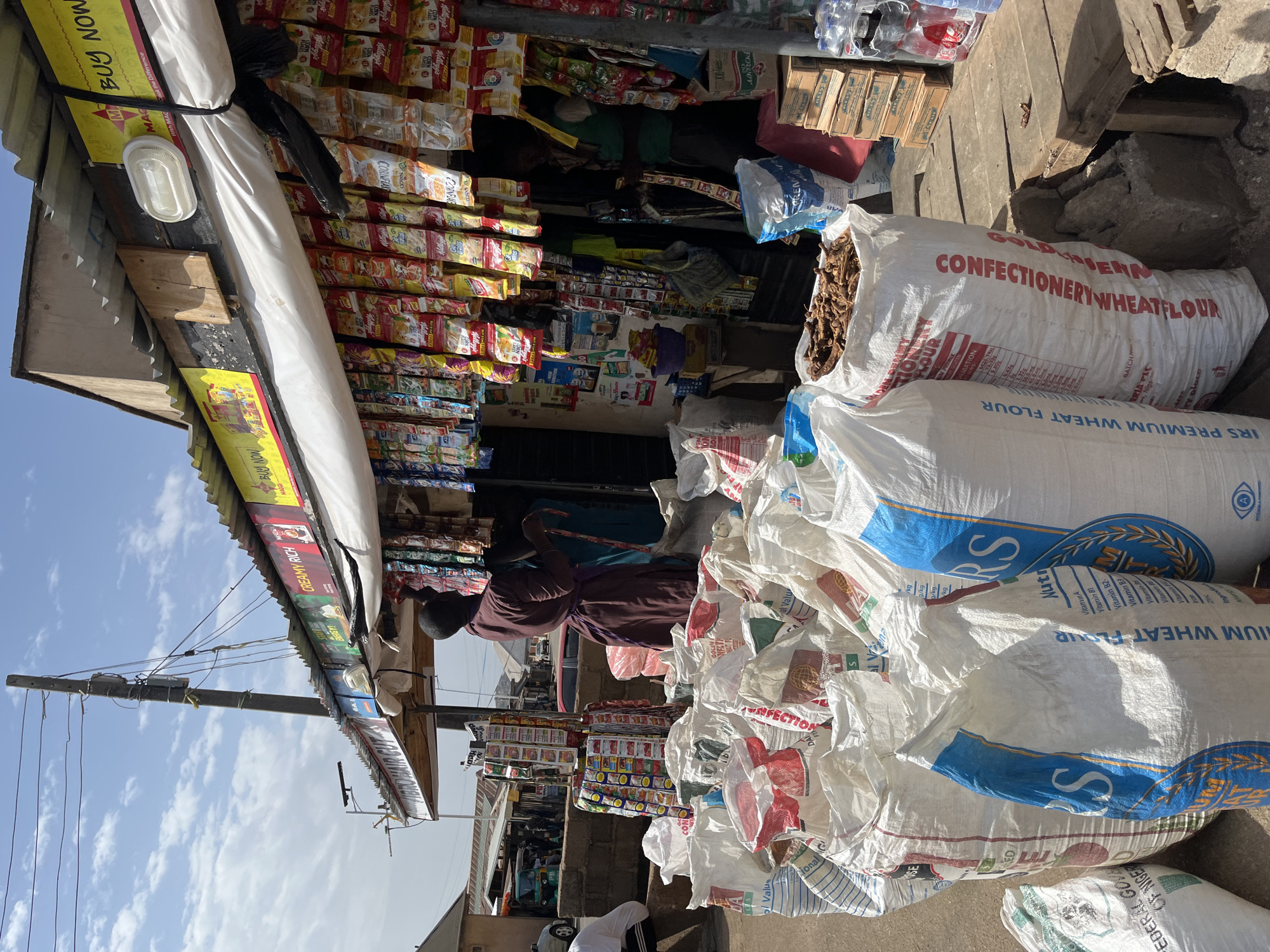 Here is a detailed description of the image:
**Scene Overview:**
The image captures a street-side market scene in what appears to be a densely populated area. The main focus is on a small open-front shop and the goods displayed around it.
**People:**
A person wearing a brown garment and a cap is present within the shop area, seemingly attending to business.
**Objects:**
* **Goods Displayed:** The shop's awning is lined with various packaged food items, including breakfast cereal boxes. Inside the shop, there are more diverse goods, including smaller packaged items, stacked densely.
* **Sacks of Wheat Flour:** A prominent feature of the image is the presence of numerous large white sacks stacked on the ground in front of the shop. The sacks are labeled with "CONFECTIONERY WHEAT FLOUR" and "IRS PREMIUM WHEAT FLOUR." One of the sacks is split open and shows a quantity of dried brown sticks.
* **Shop Structure:** The shop has a simple, corrugated iron awning and a wooden frame.
* **Power Lines:** Utility lines and a utility pole are visible in the background, suggesting an urban or semi-urban setting.
**Location (Inferred):**
Although not explicitly stated in the image, based on the items displayed in the shop and details on the bags of wheat flour ("FEDERAL GOV"), the picture could be in a local Nigerian market.
**Actions and Interactions:**
The person in the shop appears to be engaged in serving or managing the products. The sacks of flour are positioned for easy access, likely for sale.
**Environment:**
The setting appears to be outdoors, with a visible sky. The ground is a light color, suggesting a concrete or paved area. The image appears to have been rotated to the side, to better fit the aspect ratio of the image.
**In conclusion, the image depicts a small shop in a market area. It is filled with a wide variety of packaged goods, as well as large bags of wheat flour, which create a focal point in the foreground.**