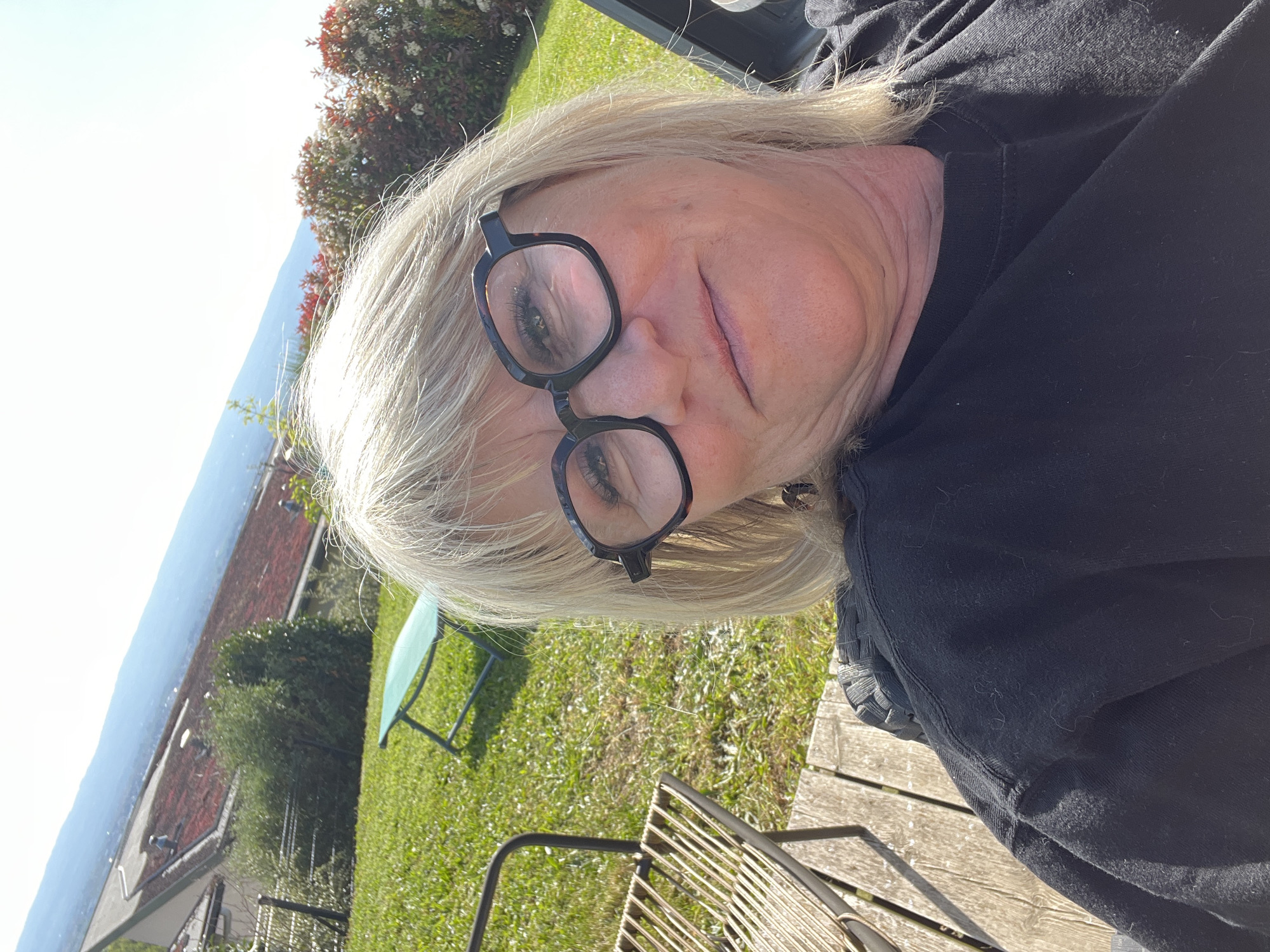 The image is a rotated selfie taken outdoors, featuring a person with blonde or light grey hair wearing dark-framed glasses. Her face, including her eyes, nose, and slightly parted lips, is visible. She wears a dark, long-sleeved top or sweater.
The setting is a sunny outdoor environment, likely a garden or yard in Chambœuf, France. Bright green grass covers the ground, and parts of outdoor furniture, including a light green metal chair and a wooden deck or patio with a metal chair, are visible. Lush bushes and trees with green leaves, some red foliage, and small white flowers frame the background.
Beyond the immediate garden, red-tiled rooftops of buildings can be seen. In the distance, a large body of water, consistent with a lake like Lac de Chaumeix, stretches towards a distant landscape of hills or mountains under a clear, bright sky. The strong sunlight indicates it is daytime, likely morning or afternoon. The person's expression is neutral, suggesting a moment of relaxation. There is no visible text in the image.