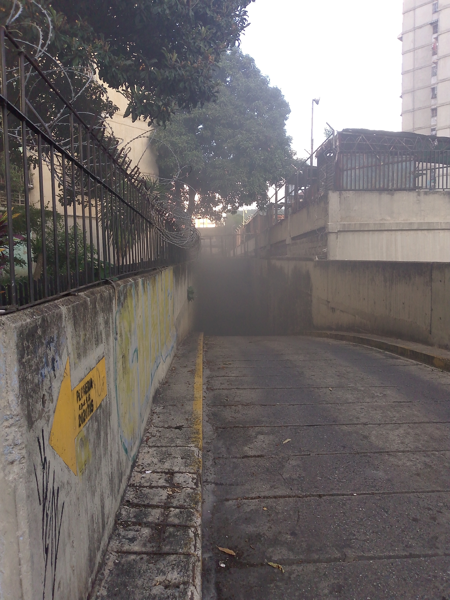 The image depicts an entrance to a dimly lit tunnel or underpass, likely part of a parking garage or a pedestrian walkway, situated in an urban environment. The area appears to be in Caracas, Venezuela, given the context provided.
In the foreground, a concrete wall is visible on the left, covered with graffiti. A yellow arrow pointing to the left is painted on this wall, with some lettering beneath it that includes "PLUMBERIA" and "20201785". Adjacent to the wall is a narrow, paved pathway with a yellow line marking its edge. Beyond this pathway is the entrance to the tunnel, which is filled with dark shadows and appears to be emanating a haze or smoke.
To the right of the tunnel entrance, a paved ramp slopes downwards into the darkness. The ramp is made of asphalt and shows signs of wear, with visible cracks and seams. The surrounding environment includes other concrete structures and a metal fence topped with barbed wire, suggesting security measures. In the background, trees with dark green foliage are present, and a multi-story building, typical of urban architecture, is partially visible against a hazy sky. The overall atmosphere is somewhat industrial and possibly neglected, with elements of urban decay such as graffiti and weathered concrete. The presence of haze or smoke in the tunnel entrance is a notable detail that could indicate various conditions, from exhaust fumes to atmospheric phenomena.
