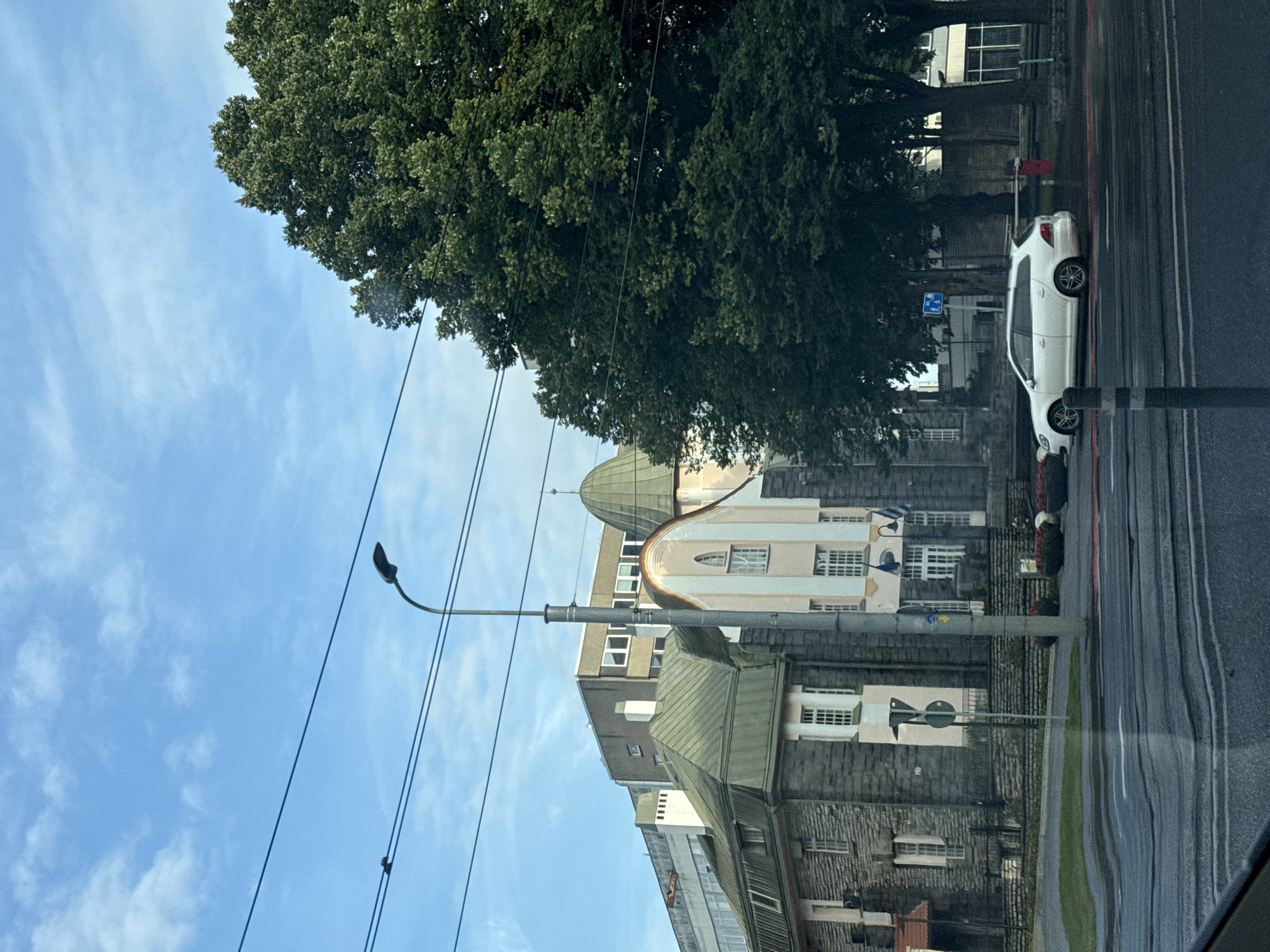 A white station wagon is parked on a wet city street in Tallinn, Estonia, near a row of buildings. The car is positioned in front of a large tree. In the background, several buildings are visible, including one with a distinctive green, domed roof and another with a stone facade. A streetlamp stands prominently in the foreground. The sky above is a clear blue with scattered clouds. The street appears to be a multi-lane road, with traffic lights and overhead wires visible. The overall scene suggests an urban setting, possibly on a slightly overcast or recently rained-on day.