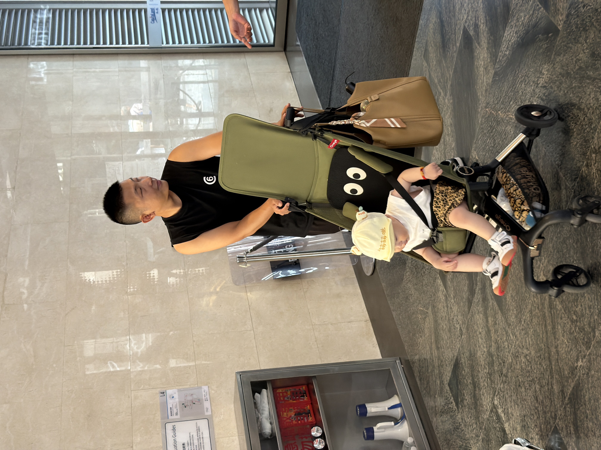 A man in a black sleeveless shirt with a white logo is standing and looking to his right. He is pushing a stroller with a baby in it. The baby is wearing a yellow hat and a white shirt with a leopard print skirt. The stroller has a black pillow with two eyes. A brown bag is hanging on the stroller. In the background, there is a wall with tiles and a metal cabinet with some items inside. A glass door is visible on the right side. The floor is tiled. The scene appears to be indoors, possibly in a lobby or public space in Yuzhong District, China.