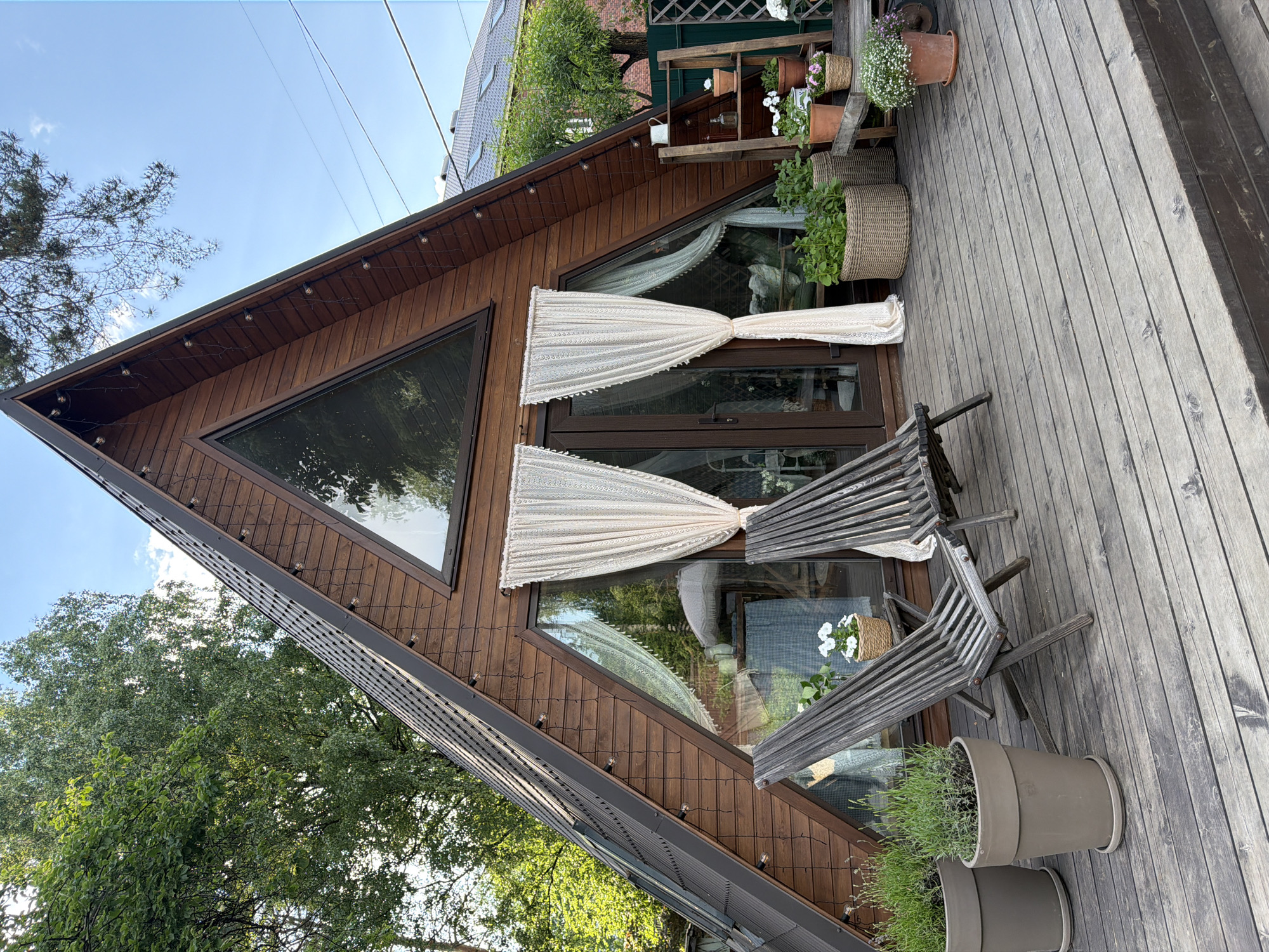 The image depicts an A-frame wooden cabin with large windows and a deck. Two empty wooden lounge chairs are placed on the deck in front of the cabin. Several potted plants are arranged on the deck and on a wooden shelving unit. The cabin is surrounded by trees and greenery, with a clear blue sky overhead. The overall scene suggests a peaceful outdoor setting.
