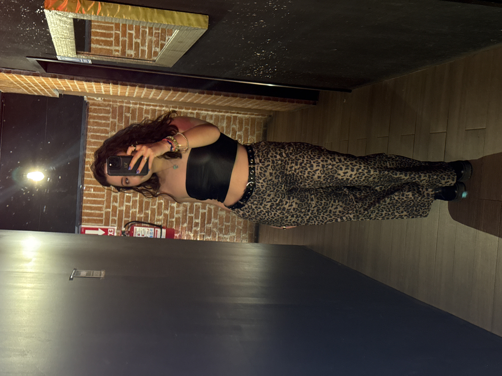 A woman stands in a room taking a picture of herself in a mirror with her smartphone. She is wearing a black strapless top, leopard print pants, and black shoes. She is also wearing a black belt with silver studs. The background includes a brick wall with a fire extinguisher. The floor is wooden and the ceiling is dark. The photo could have been taken in San Bernardino Tlaxcalancingo, Mexico, although there is not enough information in the image to determine the location specifically.