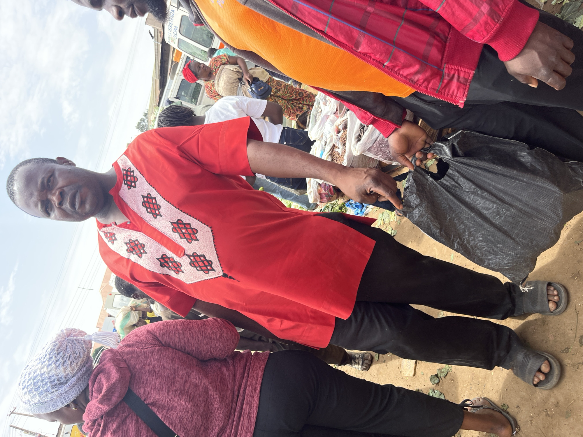 In Gura Top, Nigeria, a group of people are gathered outdoors, seemingly at a market. The scene is dominated by a man in the foreground wearing a vibrant red tunic with white and black geometric embroidery. He is looking towards the camera with a serious expression and has his hand extended, appearing to interact with a black plastic bag held by another person.
Behind him, another individual is seen from the shoulders down, wearing a pinkish-red hooded garment and a patterned white beanie. Their legs, clad in black pants, are visible along with their feet wearing sandals.
Further in the background, other people are visible, including a woman in a colorful patterned dress and a red headscarf. Some are looking away from the camera, engaged in activities related to the market. Bags of goods, possibly produce or other commodities, are scattered around, suggesting a bustling marketplace environment. A white vehicle, likely a van or bus, is partially visible in the background.
The lighting suggests it is daytime, and the weather appears to be fair. The overall atmosphere seems to be one of everyday commerce and social interaction in a Nigerian setting. No visible text is discernible in the image.