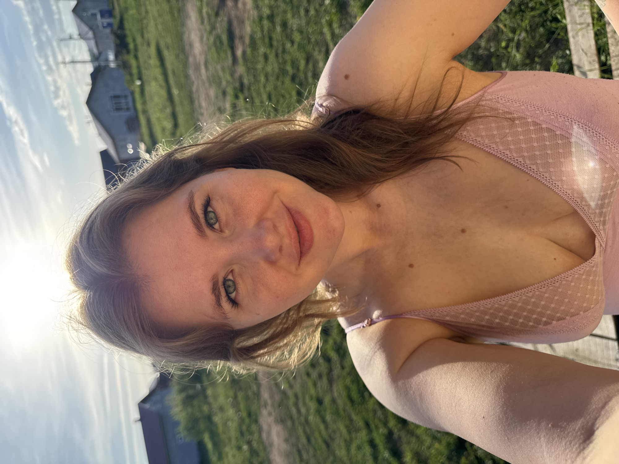 A selfie photograph shows a woman with light hair and eyes, wearing a pink top with lace detailing. She is outdoors in Ферма, Russia, with houses visible in the background under a bright sky. The foreground has green grass and the photo has warm, natural lighting.