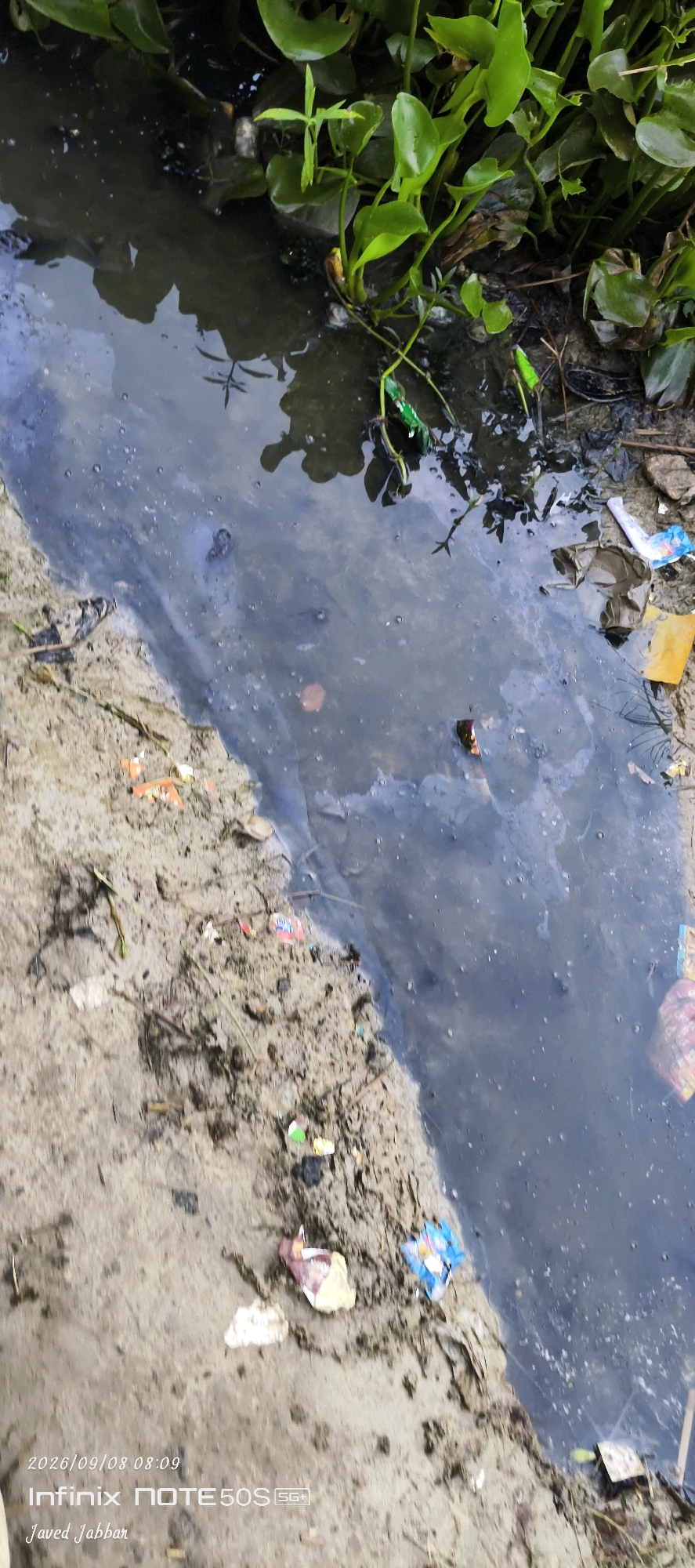 This image depicts a polluted narrow body of water, likely a ditch or small canal, running alongside a muddy bank. The water is dark and shows a visible sheen of oil or other contaminants on its surface. Scattered within the water and along the bank are various pieces of trash, including plastic wrappers and what appears to be a crumpled plastic cup. Green aquatic plants are growing along the edge of the water, contrasting with the otherwise bleak scene. The muddy bank is uneven and appears to be composed of soil and debris. Text overlays in the bottom left corner indicate the date and time as "2026/09/08 08:09" and the device as "Infinix NOTE 50S SG+", with the name "Javed Jabbar" below. The specific location of Bahera, India is mentioned in the prompt but not visible in the image itself. The scene conveys a sense of environmental neglect and pollution.