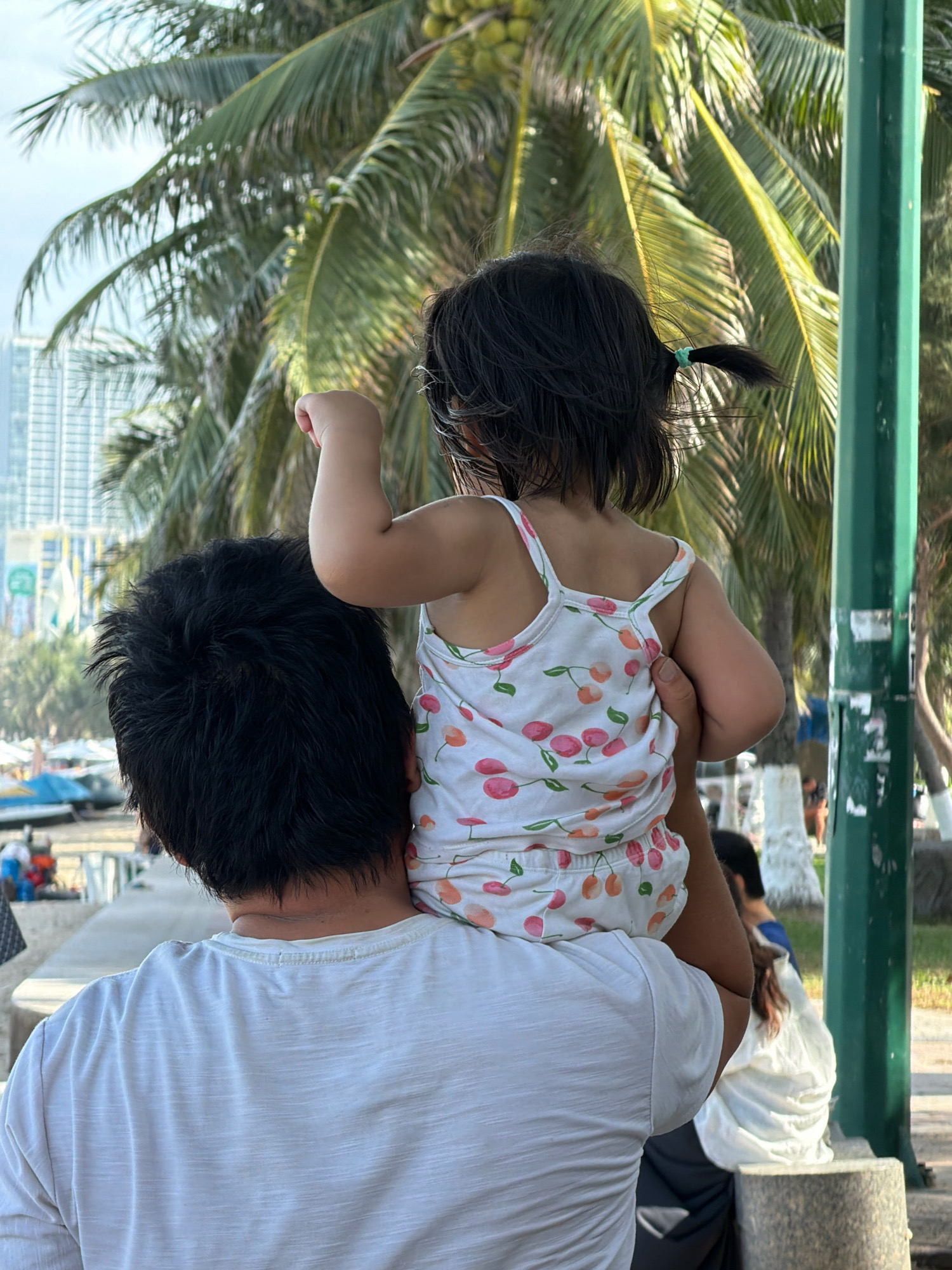 A person, seen from behind, carries a young child on their shoulders. The child, facing away from the camera, wears a white tank top with a cherry pattern. The background features palm trees, a building in the distance, and a pathway. The scene appears to be in a coastal area, likely a public space such as a park or promenade, given the presence of palm trees and what looks like a sidewalk. The lighting suggests it is daytime. The overall impression is one of a casual outing, possibly in a tropical or subtropical location. The context of Nha Trang, Vietnam, would suggest a beachside setting or a resort area.