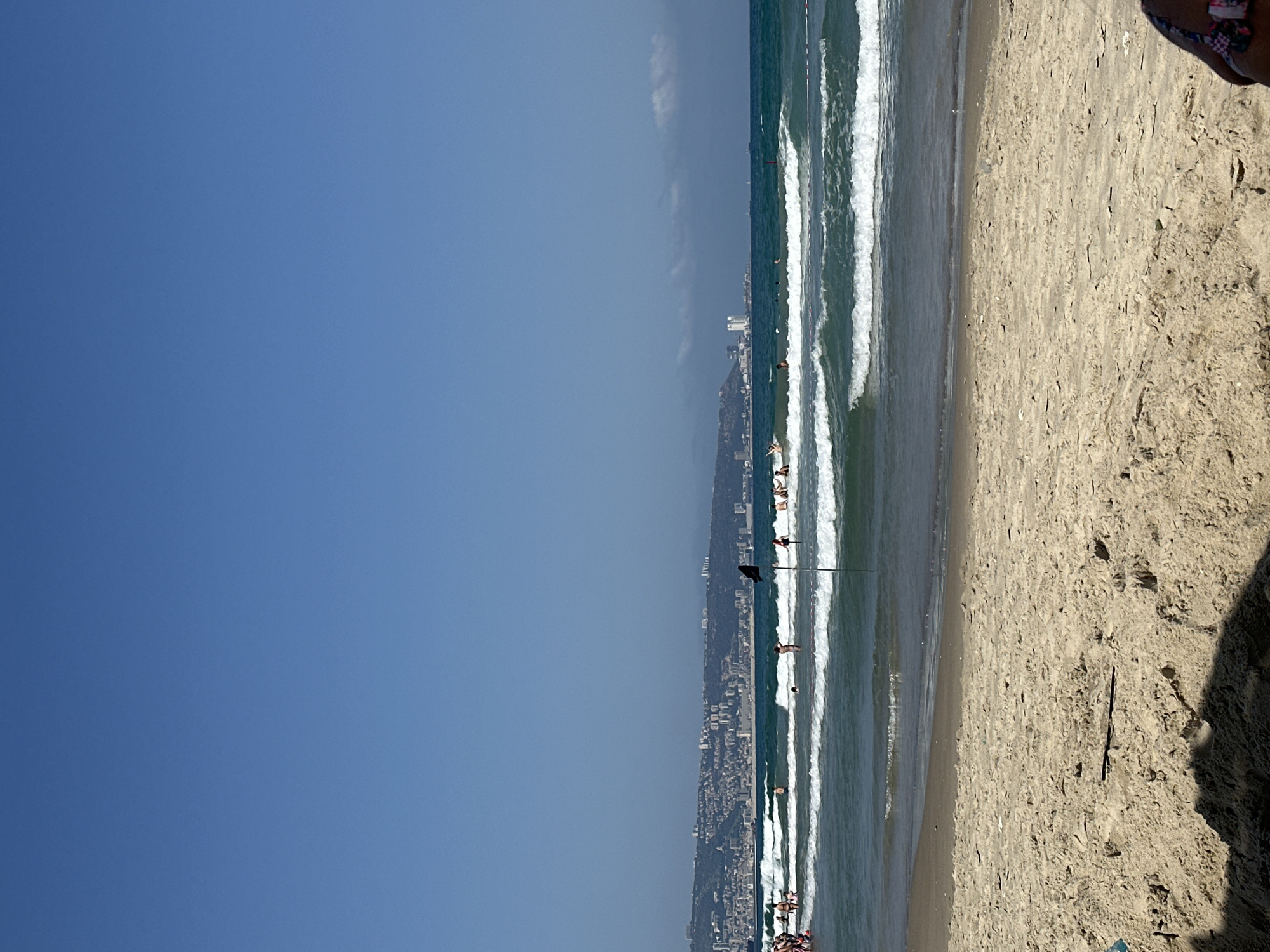 The image shows a view of the Kiryat Yam beach in Israel. The foreground is dominated by sandy beach. In the midground, the blue sea with white-capped waves is visible. Several people are seen in the water, some swimming and others standing. A black flag on a pole is planted in the sea. In the background, the city of Kiryat Yam stretches along the coastline, with buildings of varying heights. The sky above is clear and blue, with a few wispy clouds on the horizon. The overall scene depicts a sunny day at the beach with recreational activities.