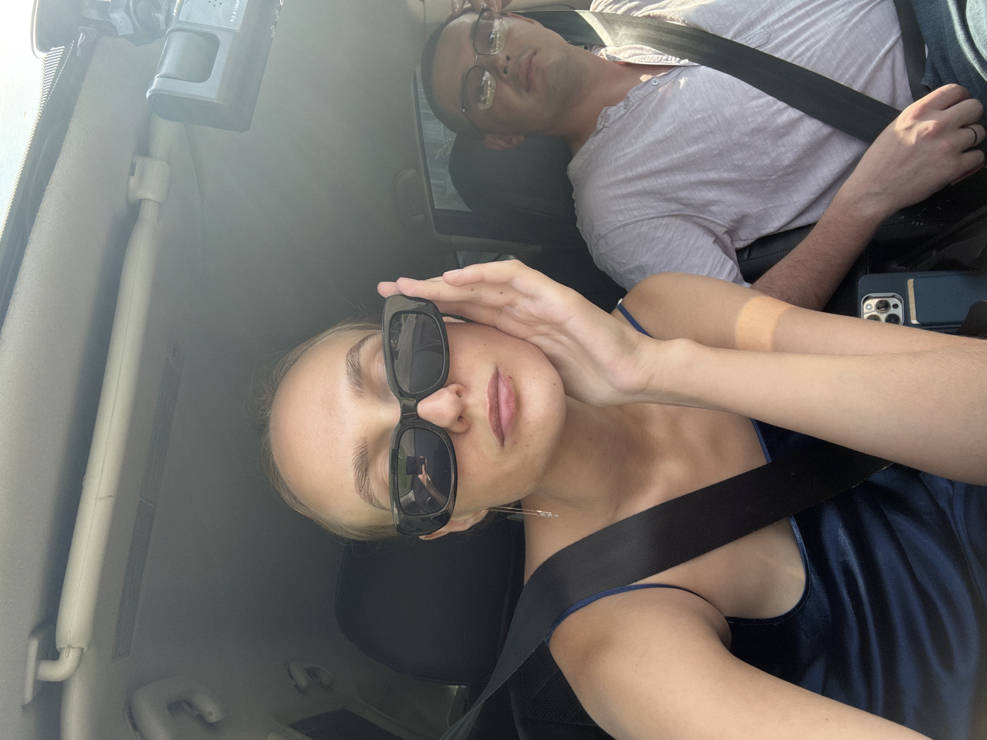 The image shows two people, a woman and a man, sitting in a car. The woman is in the foreground, wearing sunglasses and a dark blue dress, with her hand resting on her cheek. The man is in the background, wearing a light gray shirt and glasses. Both are wearing seatbelts. A dashcam is visible on the car's ceiling. The scene is consistent with being inside a vehicle, potentially in Votkinsk, Russia, given the request. The lighting suggests daytime.