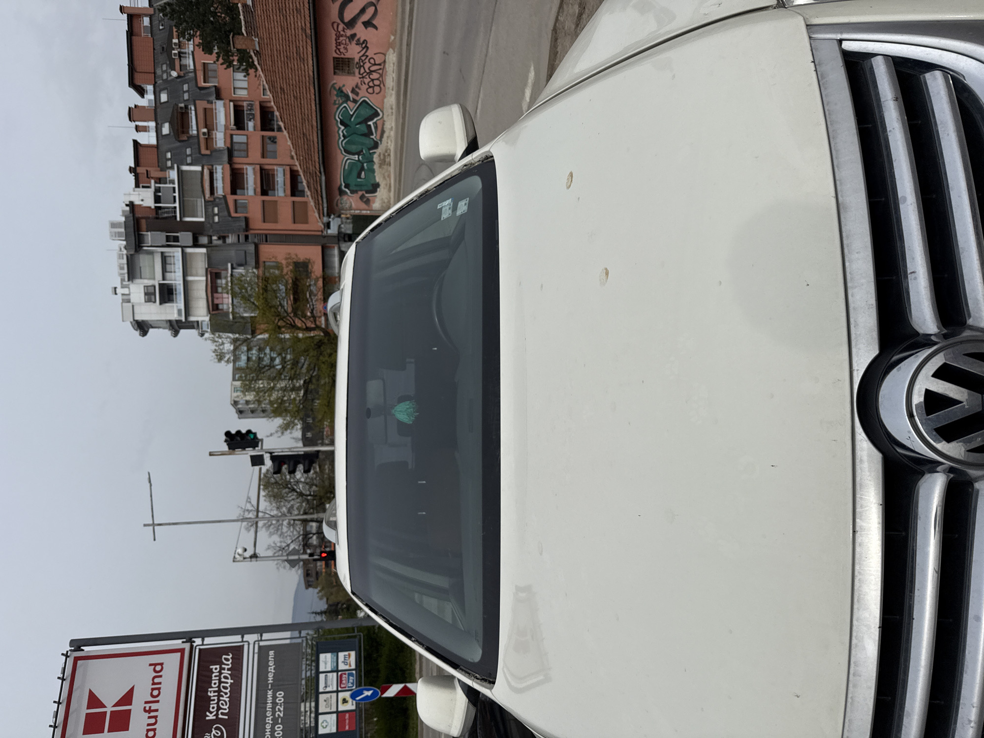 The image shows the front of a white Volkswagen car parked in front of a Kaufland supermarket in Sofia, Bulgaria. The Kaufland sign is visible, along with the supermarket's bakery section sign. The building in the background has a red roof and colorful walls with graffiti. Traffic lights are visible in the distance.