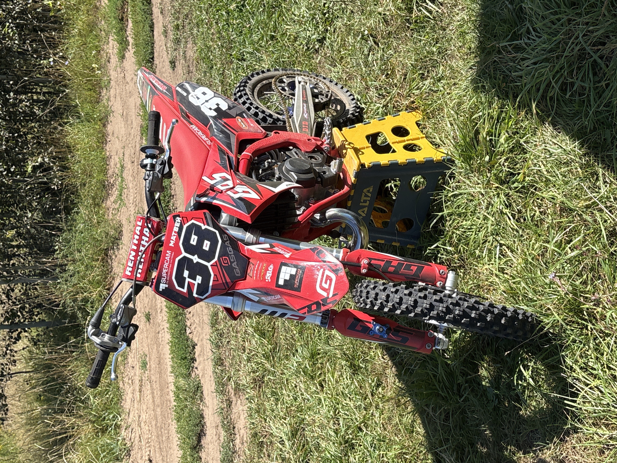 A red and black dirt bike, number 36, is displayed on a yellow stand on a grassy area next to a dirt track. The dirt bike has "GASGAS" branding and various sponsor decals. The background consists of dry grass and trees. The scene appears to be outdoors on a sunny day. There are no people visible in the image. There is no information or visual evidence to suggest the location is Votkinsk, Russia.
