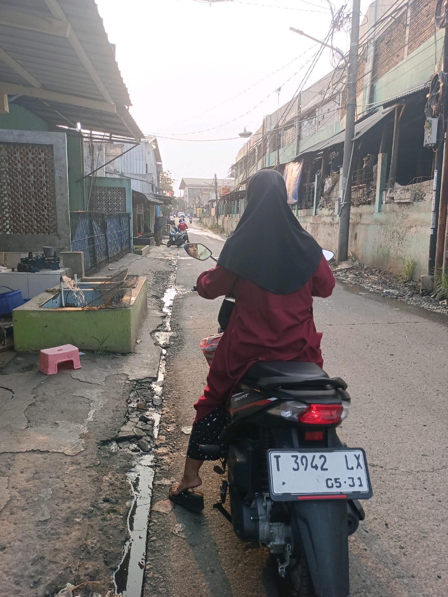 A medium shot captures a person from behind, riding a black scooter down a narrow, paved street in Karawang, Indonesia. The rider, wearing a dark hijab and a maroon tunic over patterned pants, is partially obscuring the scooter's license plate, which reads "T 3942 LX 05-31". The street is lined with buildings constructed from various materials, including corrugated metal and brick, some with green paint and others appearing weathered. In the foreground on the left, there's a cracked concrete sidewalk with a green concrete basin filled with water and a net, and a small pink plastic stool. The overall environment suggests a working-class urban neighborhood with visible signs of wear and tear, including debris along the roadside and damaged pavement. The sky is bright and hazy, indicating it is daytime, likely late afternoon due to the warm light.
