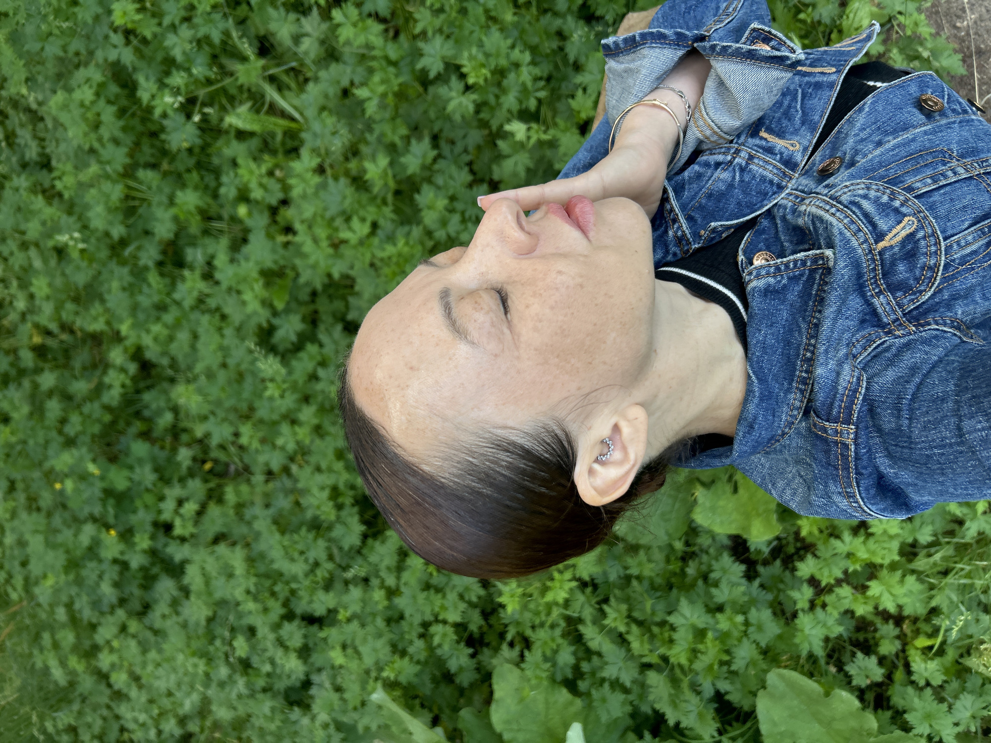 A woman with freckles and closed eyes is seen in profile, her right hand touching her face, as she stands outdoors in front of a dense green foliage background. She is wearing a denim jacket over a black shirt. Her hair is pulled back, and she has jewelry on her ear and wrist. The image does not contain information indicating the location in the city of Chaikovsky, Russia.