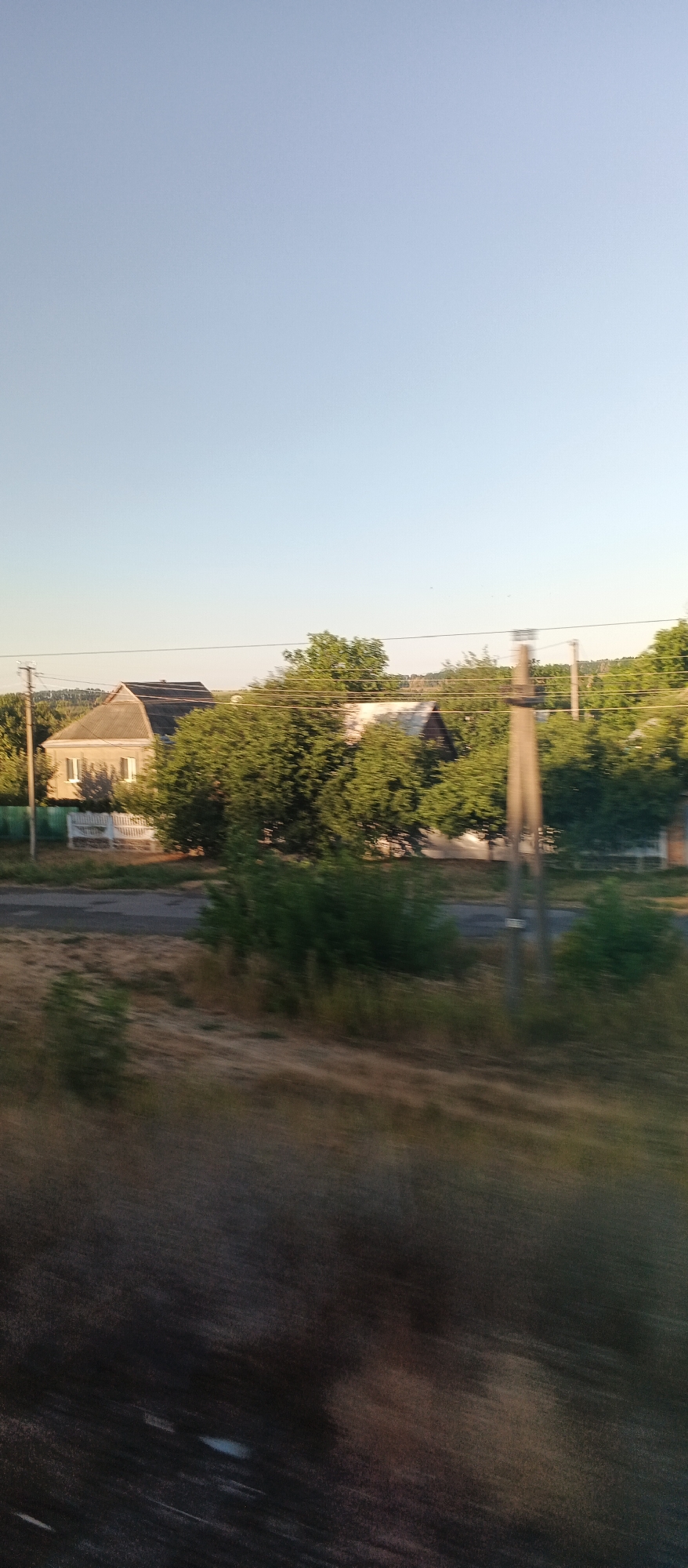 The image shows a rural landscape under a clear sky, likely in Khlystunivka, Ukraine. In the middle ground, a small, light-colored house with a gray roof is visible, partially obscured by lush green trees. A white picket fence runs in front of the house. Further to the right, another house with a darker roof is partially visible. Several tall, bare trees are interspersed with leafy trees, suggesting a mix of deciduous and perhaps evergreen species. A utility pole stands prominently in the mid-right of the frame, with power lines running horizontally across the scene. The foreground is blurred and shows dry, grassy terrain, indicating a motion blur, possibly from a vehicle. The overall impression is of a quiet, rural setting during daylight, likely late afternoon or early evening given the warm lighting.