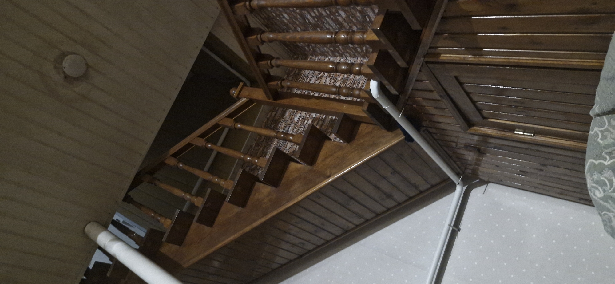 The image shows a wooden staircase inside a building, possibly a house. The staircase is made of dark polished wood and has carved balusters. The walls are paneled with wood, and there is a white pipe running along the wall. The ceiling is also paneled with wood. The overall impression is one of a rustic interior. There are no people visible in the image. The location is specified as Ordynskoye, Russia.