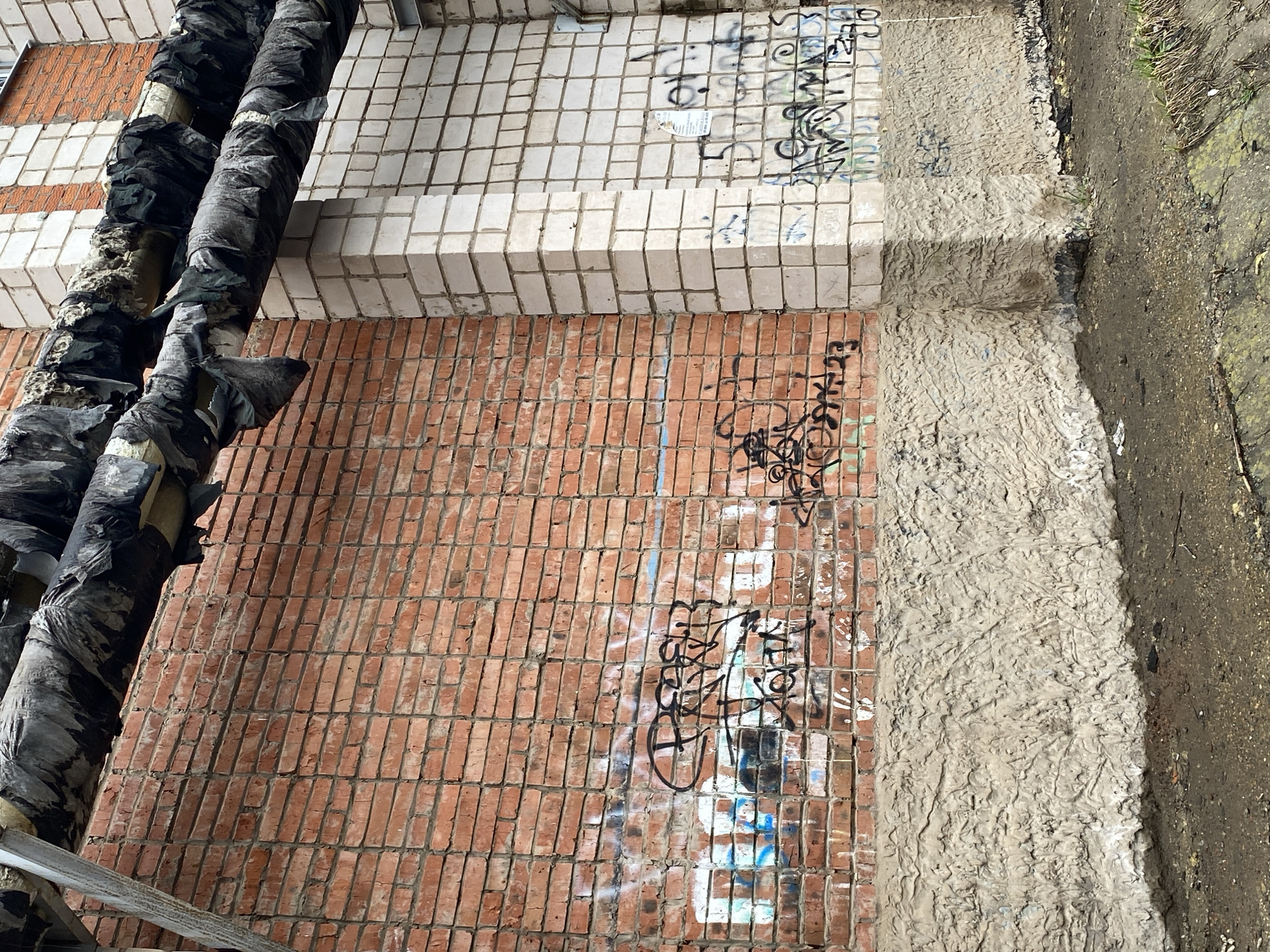 The image captures an angled, downward-looking perspective of a neglected exterior urban environment in Votkinsk, Russia.
The scene is dominated by a wall section constructed from three distinct materials. In the upper left, there is a section of a building facade covered in small, light-colored (likely white or off-white) square tiles, which are dirty and adorned with black spray-painted graffiti. Below this, a red-orange brick wall, featuring vertically oriented rectangular bricks, forms another part of the building's exterior, also heavily covered in black and some faded white/blue spray paint graffiti. A distinct concrete ledge or step separates these two wall sections, running horizontally across the image's middle.
Two large, insulated pipes, wrapped in a torn and degraded black, tarp-like material revealing lighter insulation underneath, run diagonally along the left side of the image, obscuring part of both the tiled and brick walls.
To the right of the wall sections, the ground transitions to a roughly textured, light grey concrete-like surface, which then gives way to a dark, muddy, and dirty patch of earth with sparse green vegetation along the far right edge.
No people or direct activities are visible. The time of day appears to be daylight, possibly overcast, given the lack of strong shadows. The muddy ground suggests a damp environment or recent precipitation. The overall impression is one of urban decay and utilitarian infrastructure, common in older industrial or residential areas. The graffiti, consisting of stylized tags and scrawls, is largely illegible.