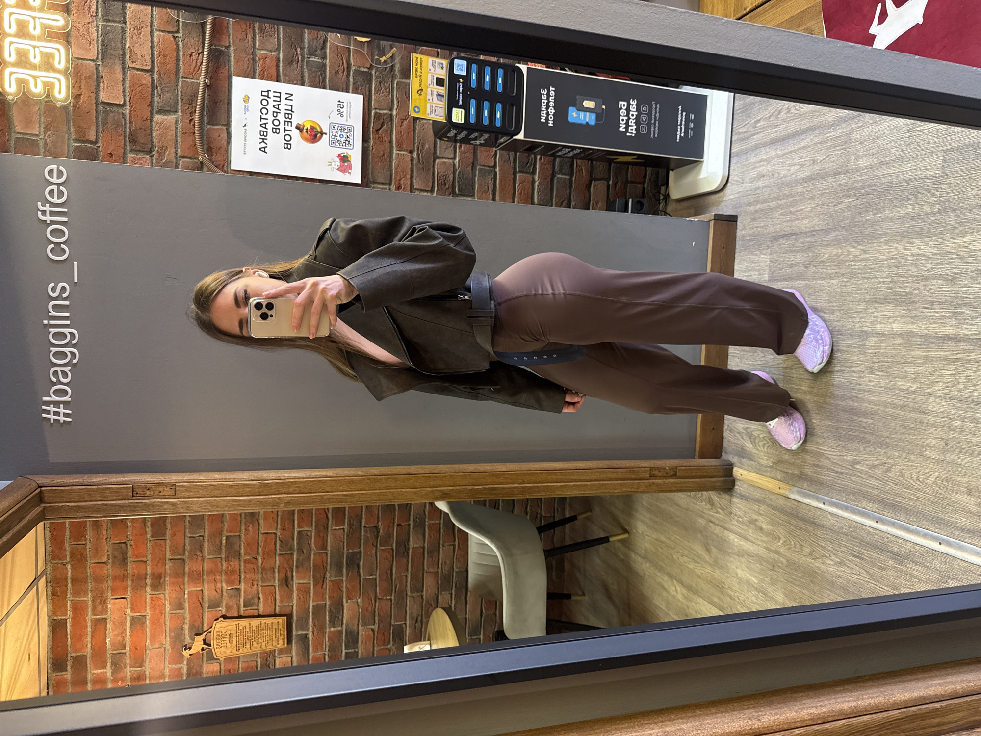 A woman is taking a selfie in a mirror located in what appears to be a coffee shop or café called "baggins coffee." The location is in Saint Petersburg, Russia, based on the Cyrillic text on the signage and advertising materials visible.
The woman is wearing a brown jacket, brown pants, and lavender shoes. She is holding her phone up to take a picture of herself in the mirror.
The coffee shop has brick walls, and there are various signs and advertisements on the walls in Russian. The signage indicates information related to coffee and phone charging stations. There is also furniture visible in the mirror's reflection, including a chair and a table. The floor is made of wood.