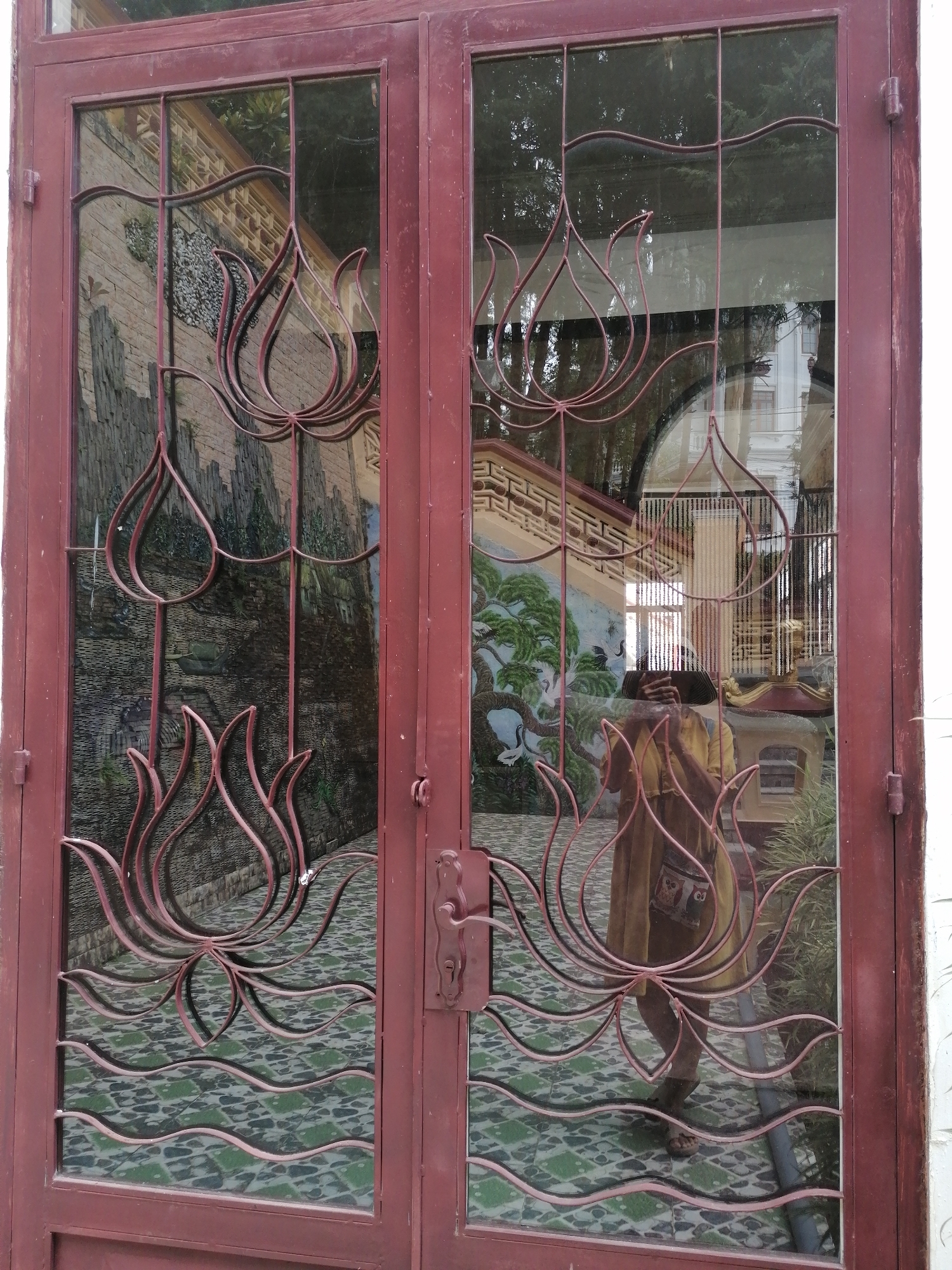 This image shows a double-door with ornate ironwork featuring lotus flower designs. The doors are made of dark red metal frames with glass panels. The glass panels reveal reflections of a tiled floor with green and white geometric patterns, and a mural or artwork depicting a rocky landscape with vegetation on the left door, and a scene with trees, birds, and architectural elements on the right door. A person is visible in the reflection on the right door, holding a phone and taking a selfie. The person is wearing a yellow dress and sandals. The overall impression is that these are exterior doors, possibly to a building or garden, with decorative elements that suggest a cultural or artistic theme. The specific location in Da Lat, Vietnam, is not directly identifiable from the image itself, but the style of the artwork and decorative elements might be characteristic of Vietnamese architecture or gardens.