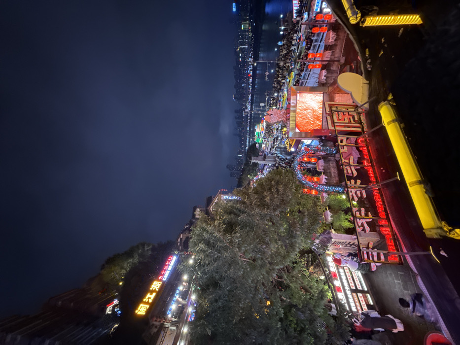 This image captures a vibrant nocturnal scene within Yuzhong District, China. The primary subject is a multi-tiered architectural complex built into a steep hillside, overlooking a wide river. The sky above is dark and cloudy.
In the mid-ground, across the river, a distant city skyline is visible, brightly illuminated. Along the near riverbank, a large crowd of people congregates on a promenade. Several boats or floating structures, also illuminated, are present on the river. A prominent, large LED screen near the river's edge displays a dynamic red-orange visual. To its left, a light-adorned archway with red lanterns provides access to a densely populated area. Additional red lanterns are spaced along the riverfront railing.
Closer to the viewer, the hillside complex features traditional Chinese-style buildings integrated into the terrain and surrounded by dense trees. These structures are extensively illuminated with colorful lights and neon signs; one prominent sign displays the Chinese characters "闯江湖" (Chuǎng Jiānghú). Multiple levels of walkways and platforms are visible, populated by numerous individuals. On one lower walkway, a human-sized Santa Claus figure stands next to an illuminated sign containing Chinese characters and numerical text, which may indicate pricing information. Red-lit railings and decorative elements are prevalent throughout the complex. The immediate foreground includes a partial view of a yellow and dark-colored barrier, indicating the observer's elevated position.