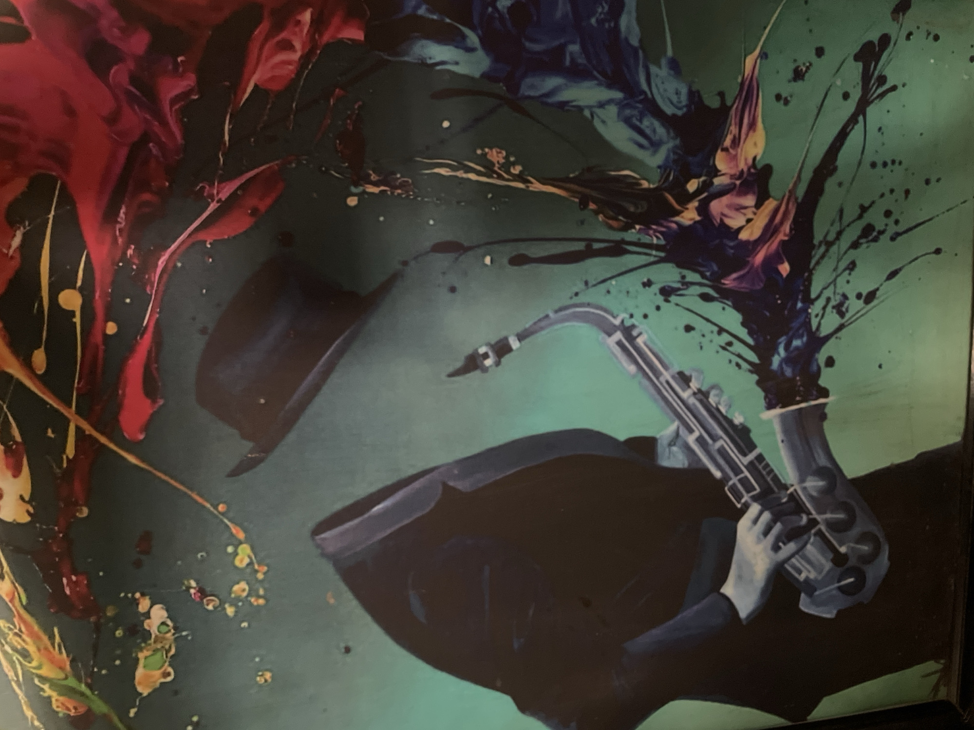 This image is an artwork depicting a figure in a black suit and hat, playing a saxophone. The figure's head is obscured by the hat, and the artwork features abstract splashes of red, blue, yellow, and green paint. There is no discernible location in the city of Abuja, Nigeria within the image.