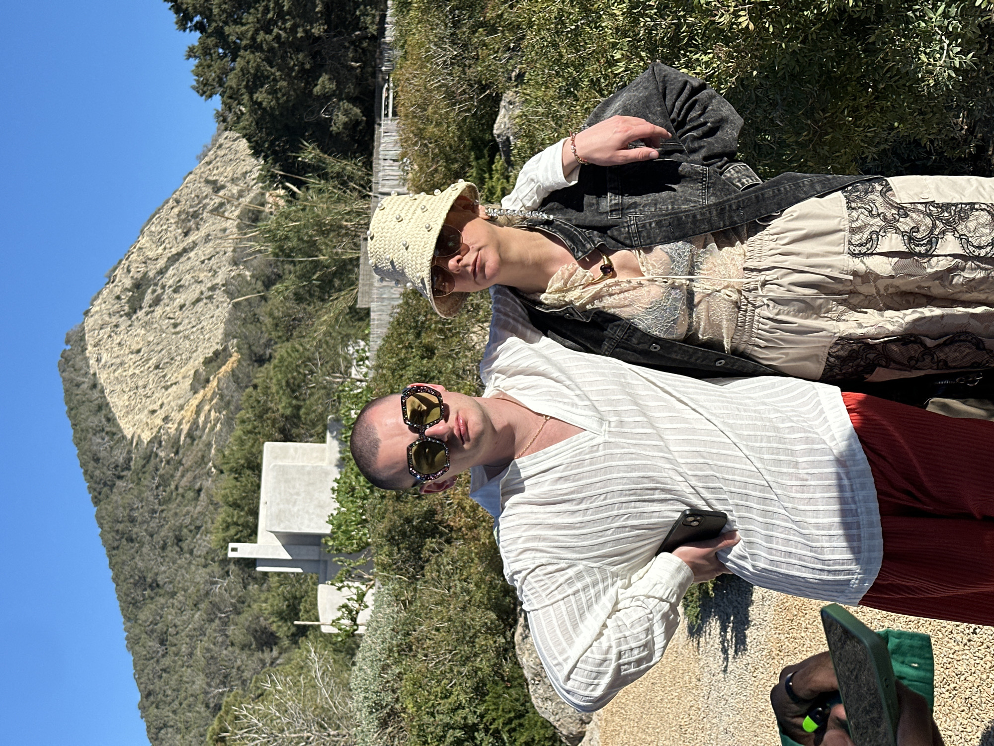 Two fair-skinned individuals pose outdoors in Sant Josep de sa Talaia, Spain. The person in the foreground wears a white, vertically-striped long-sleeved shirt, reddish-brown pants, and ornate sunglasses, holding a phone in their right hand. They are facing forward and slightly to the right of the frame. The second person stands behind, wearing a light-colored dress with lace details, a dark denim jacket, sunglasses, and a light-colored sun hat embellished with pearls. A mountain and trees are visible in the background, with a concrete structure and clear blue sky also present. The location appears to be a rocky, somewhat arid terrain. The angle of the photo is slightly rotated.