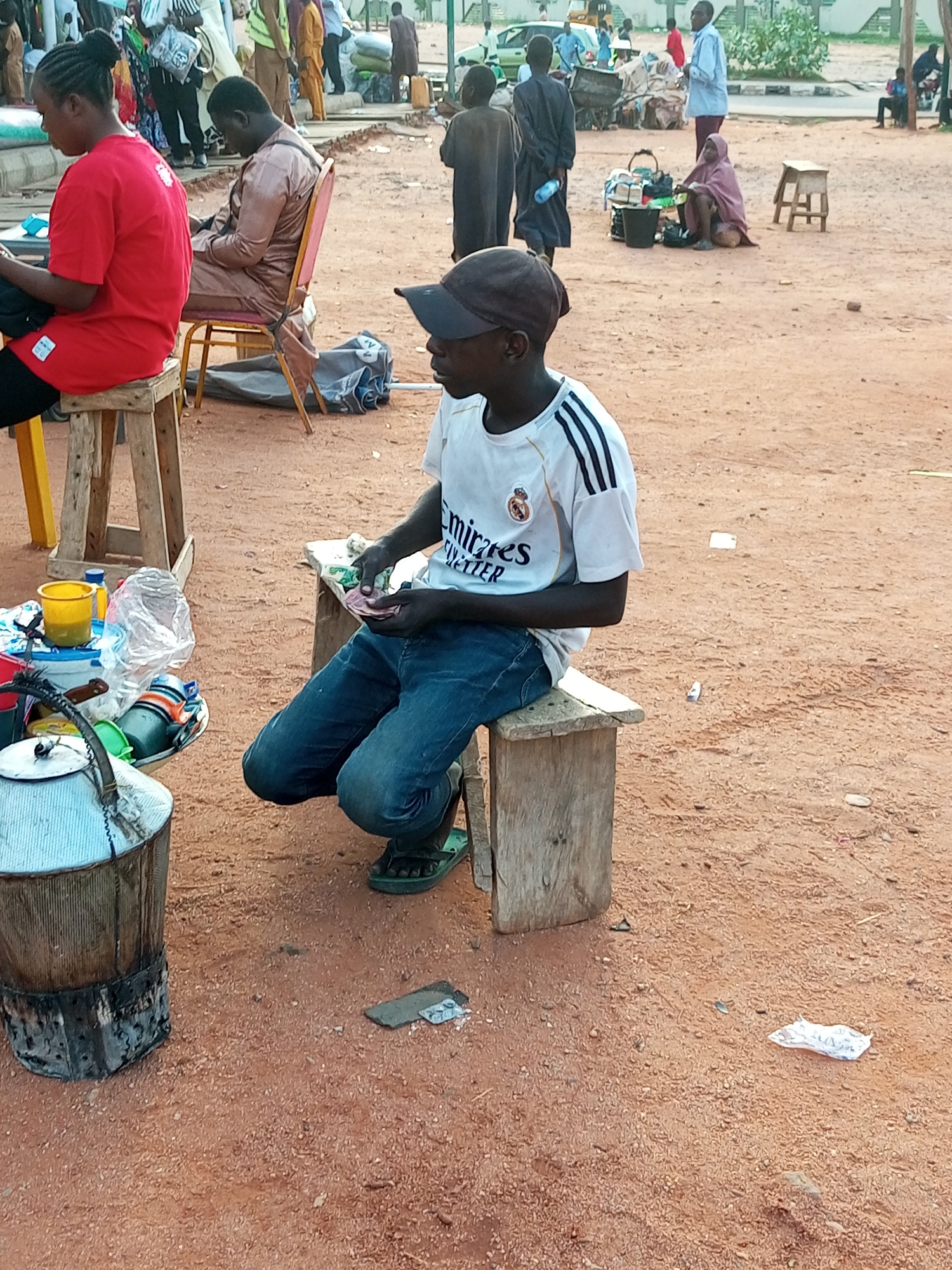 In the city of Gombe, Nigeria, a young boy wearing a white t-shirt with black stripes on the sleeves and a dark baseball cap is seated on a wooden stool in an outdoor marketplace. He is holding a wad of currency in his hands and appears to be counting it. He is wearing blue jeans and dark sandals. To his left, on a table, are various items including cups, a plastic bag, and a large metallic container. Behind him, other people and market activities are visible in the background. The ground is covered in reddish-brown dirt, typical of an arid or semi-arid environment.