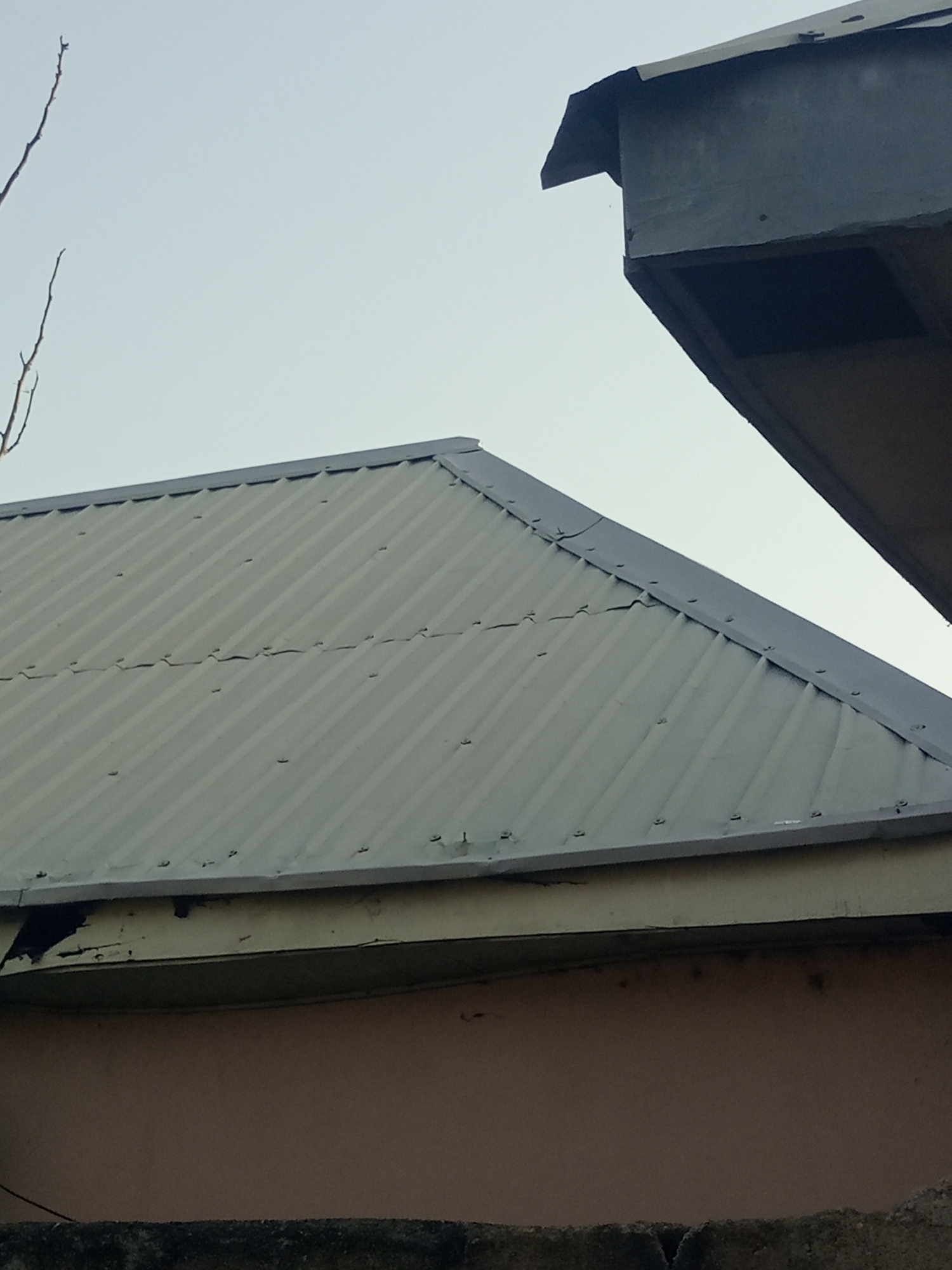 The image shows the upper section of two buildings with corrugated metal roofs, viewed from a low angle against a pale sky. The primary focus is on a large, gray corrugated metal roof that slopes downwards from left to right, its surface marked by visible seams and evenly spaced fasteners. A lighter-colored fascia board runs along the edge of this roof, beneath which a pinkish-tan wall is partially visible.
To the right and slightly higher, a portion of another structure is visible. This structure has a dark gray, angular roof overhang that juts out. The underside of this overhang reveals a dark recess, possibly an opening or ventilation. A thin, bare tree branch extends into the upper left corner of the frame. The overall scene appears to be an outdoor urban environment in Jalingo, Nigeria, likely residential or commercial, given the building structures. There are no people or animals visible in the frame, and no discernible actions are taking place. The lighting suggests it is daytime, but the sky is overcast or hazy.
