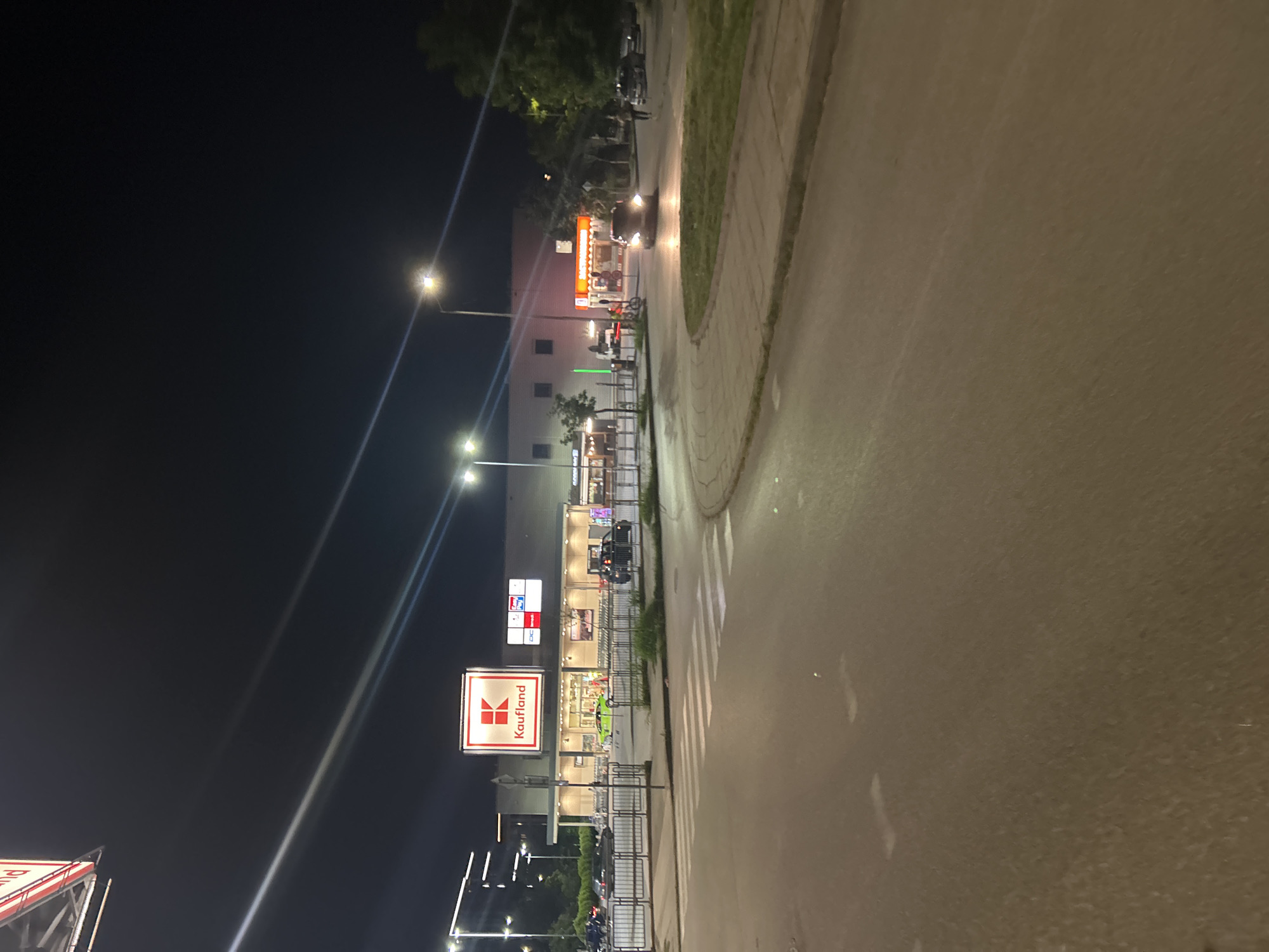 This nocturnal image captures a commercial street scene, likely in an urban or suburban area. The perspective is slightly elevated, looking down along a curving road and a commercial complex.
On the left, a multi-story building houses retail establishments, brightly illuminated by external lights and prominent signage. A large, vertically oriented, illuminated sign for "Kaufland" is clearly visible, featuring its distinctive red logo with white text. Above it, another smaller sign displays "OOC". Several indistinct human figures are present near the building, some appearing to stand or walk, and possibly some near bicycles, though details are obscured by darkness and distance. A dark metal fence separates the building's frontage from the public sidewalk.
To the right, an asphalt road stretches into the distance, appearing reflective under the artificial lights. A car with its headlights on is driving towards the viewer in the middle ground. A concrete sidewalk with a curb borders the road, and faint pedestrian crosswalk markings are visible on the asphalt. Bright streetlights line the road and foreground, creating distinct beams of light and lens flares against the dark night sky. Some trees and other vegetation are visible in the background and along the street. The scene depicts a typical evening in a commercial district, consistent with a location in Plovdiv, Bulgaria, where Kaufland supermarkets are common.
