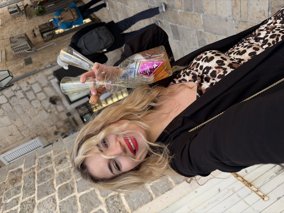 A blonde female is positioned in the foreground of a narrow, stone-paved alleyway within the Old Town of Budva, Montenegro. She is smiling directly at the camera, wearing red lipstick, a black blazer, and a leopard-print top. Her right hand holds an ornate glass decanter containing an amber-colored liquid, labeled "FABRIQUE VICTOR & JORDAN," along with two empty champagne flutes. One flute has a cork inserted into its opening. A thin, gold-colored chain crosses her chest. The alley features rough-hewn stone walls on the left and a building with white louvered shutters and an external air conditioning unit on the right. In the background, two individuals are walking away from the camera: one wearing a dark jacket and a light cap with a dark backpack, and another in a blue jacket with a tan backpack. An overhead light fixture is visible above the primary subject.