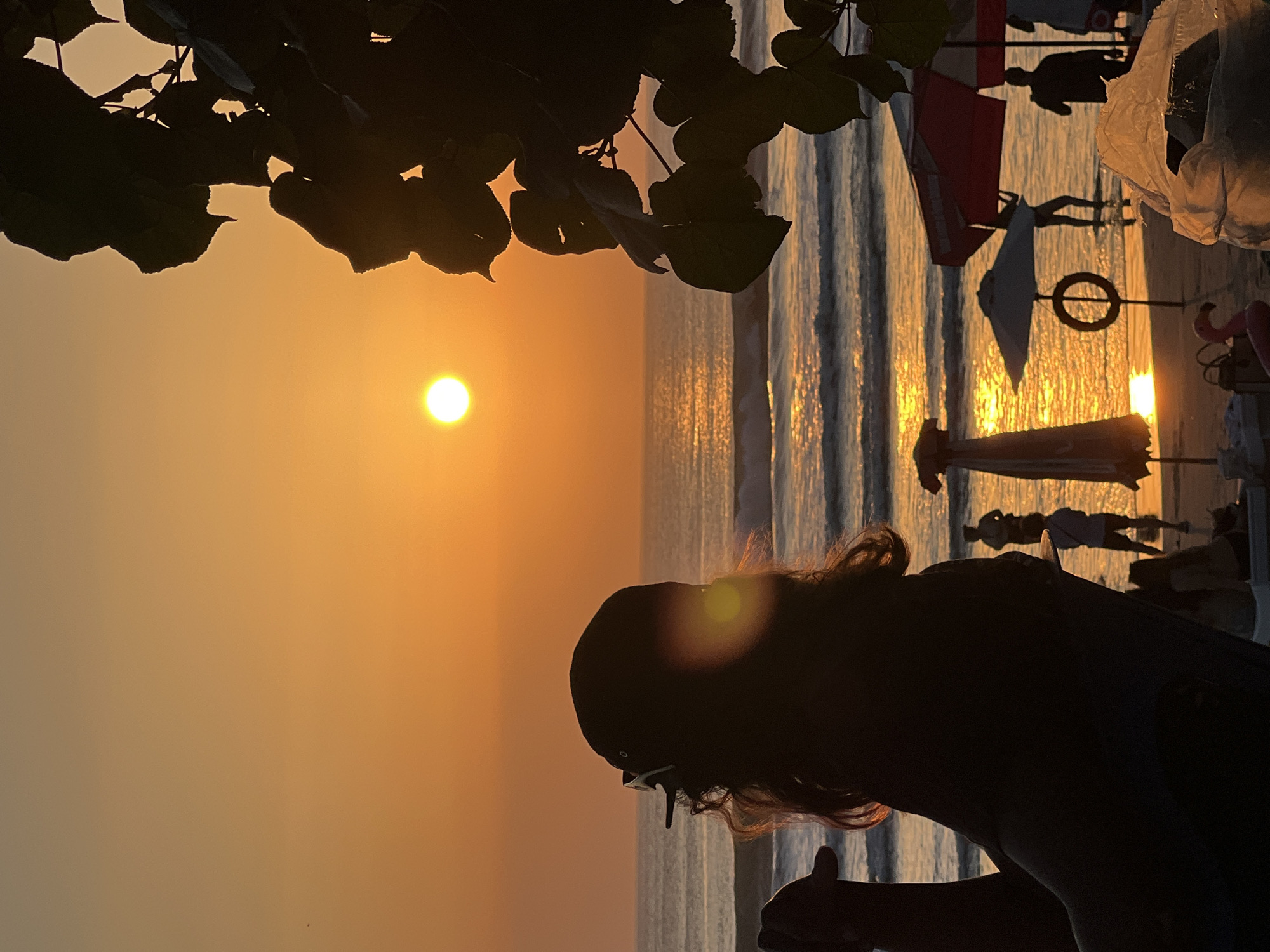 The image depicts a beach scene in Kuta, Indonesia, during sunset. The sun, a bright orb, is low on the horizon, casting a warm orange glow across the sky and water. The ocean has gentle waves reflecting the sunlight. In the foreground, silhouetted against the sunset, is a person wearing a baseball cap, their back to the camera, looking out at the sea. To the right, the dark silhouette of tree leaves frames the scene. Further down the beach, other people and beach umbrellas are visible, also silhouetted. A life buoy hangs from a stand, and a pink flamingo float is also present. The overall atmosphere is tranquil, with the natural beauty of the sunset dominating the view.
