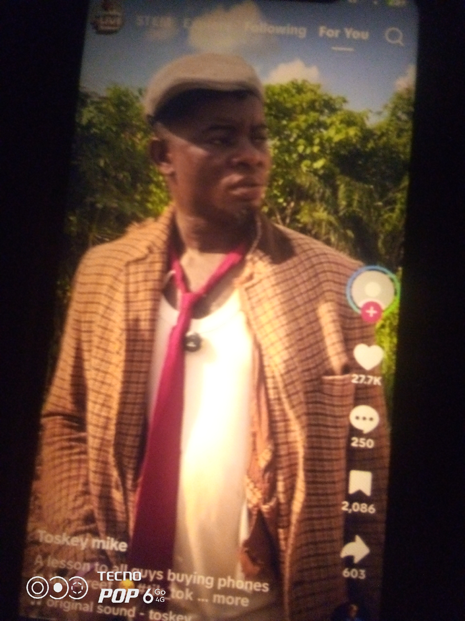 A full-shot photograph taken from a mobile device shows a man in the center of the frame. He is wearing a tan checkered jacket over a white t-shirt and a pink tie. He also wears a light-colored flat cap. The background appears to be a natural setting with lush green foliage and a blue sky with some clouds.
On the right side of the screen, there are social media icons with associated numbers: a user icon with a plus sign, a heart icon with "27.7K" below it, a comment icon with "250" below it, a bookmark icon with "2,086" below it, and an arrow icon with "603" below it.
In the bottom left corner, text is visible, including "Toskey mike," "A lesson to all guys buying phones," and "original sound - toskey." There is also a logo that appears to be for "TECNO POP 6 Go 4G."
The image quality is low, with a blurry and pixelated appearance. The lighting is uneven, and there is a strong glare from the device screen. The overall impression is that this is a screenshot or photo of a social media video playing on a phone. The exact location of Howayi, Nigeria, is not visually discernible from the image itself, but the text suggests a Nigerian context.