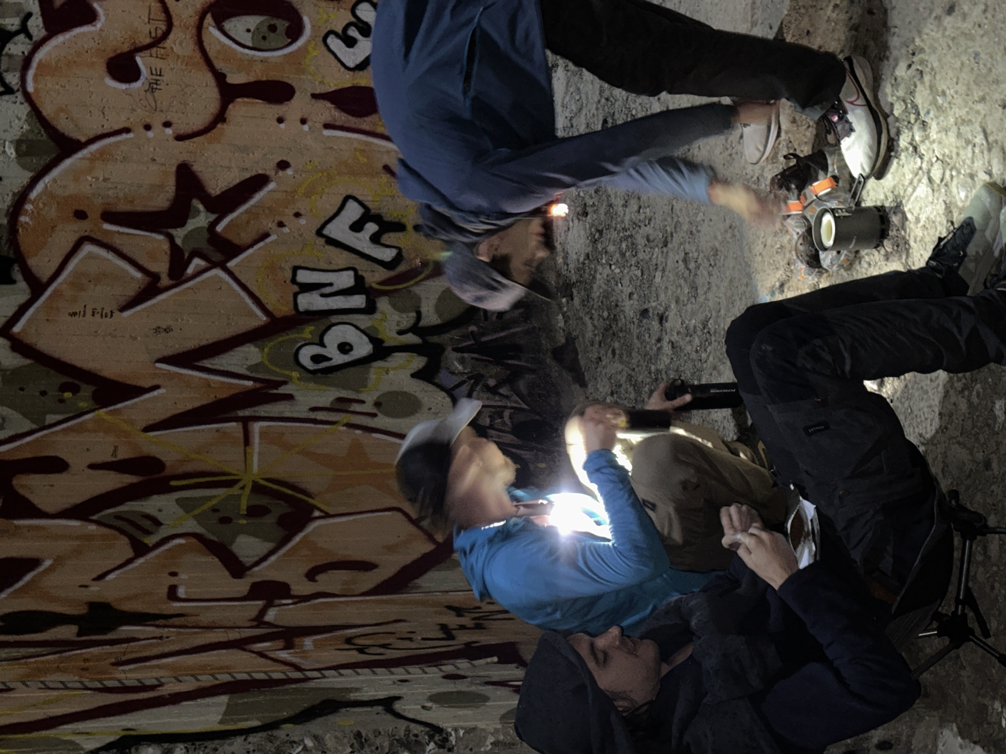A group of people stand in an urban environment, possibly a tunnel or underpass, marked with graffiti on the walls. The people are dressed in casual attire, and some appear to be using flashlights or headlamps to illuminate the area. One person is holding a camera or phone, seemingly taking a picture of the graffiti. A tripod is visible, suggesting they may be documenting the graffiti. There is evidence of concrete and stone in the environment.