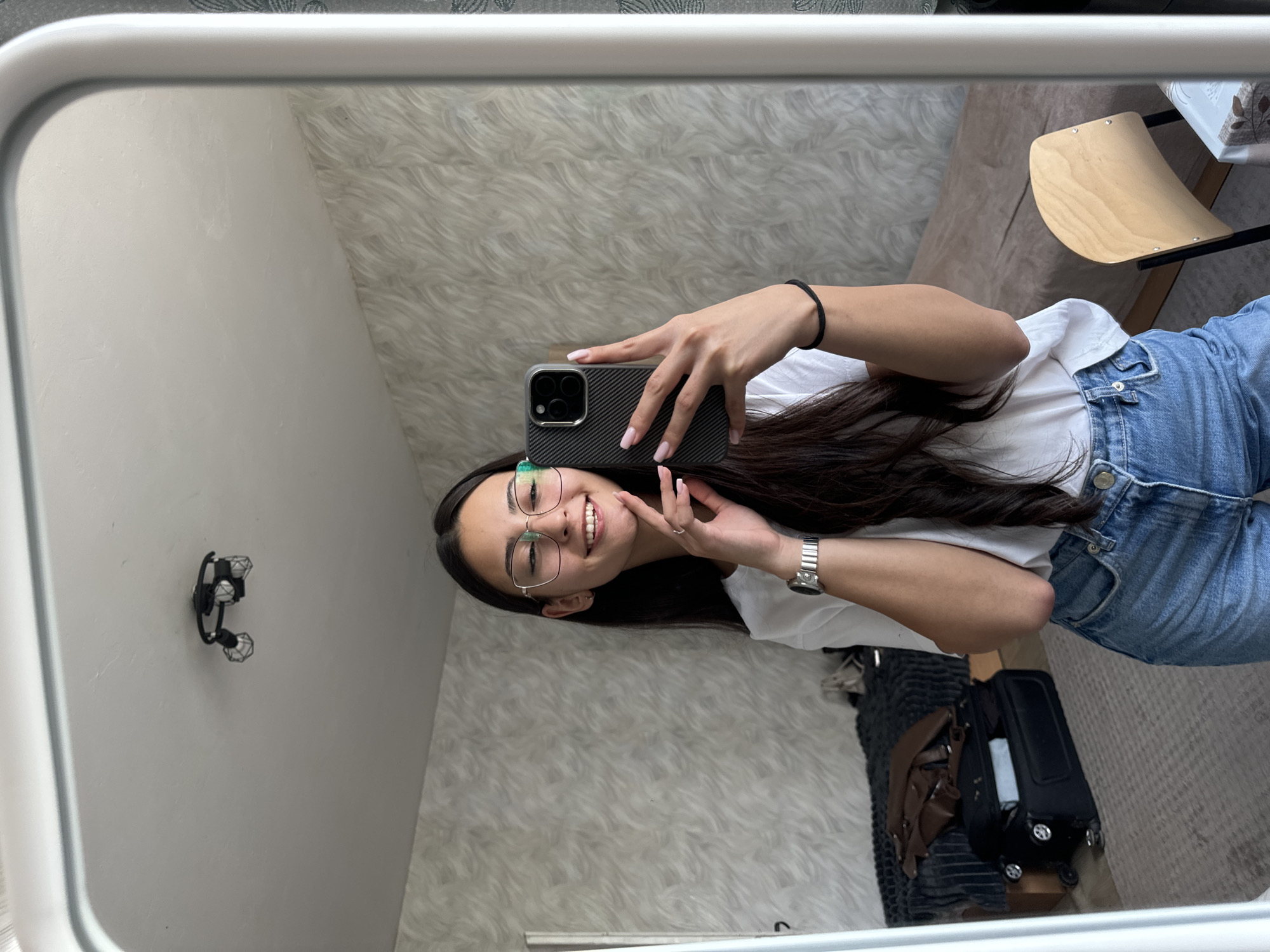 This media file is a mirror selfie, rotated 90 degrees clockwise, featuring a young woman smiling broadly. She is positioned horizontally in the frame, wearing clear-rimmed glasses, a white t-shirt, and blue jeans. She has long, dark hair and light pink nail polish.
Her right hand holds a smartphone with a dark, textured case, seemingly a carbon fiber pattern, towards the mirror to capture the selfie. A black hair tie is on her right wrist, and a silver watch is on her left wrist. Her left hand is raised, with her index finger and thumb gently touching her chin, completing a casual pose.
The reflection in the mirror reveals parts of an indoor room. Directly behind the woman's head and upper body is a textured wall or wallpaper featuring a swirling, intricate pattern in white and cream tones. In the upper left corner of the reflected room (relative to the rotated image), a black sconce light fixture with two small, geometric-shaped light shades is mounted on a plain light grey wall. To the right of the woman, a light brown wooden chair seat with black metal legs is partially visible. Further right, a black wheeled suitcase containing some light-colored items, along with a brown bag or purse, rests on what appears to be a grey rug or carpet. The mirror itself is framed by a thick, light grey border.
The overall impression is casual and cheerful. The provided context indicates the location is Sofia, Bulgaria. No visible text is discernible in the image.