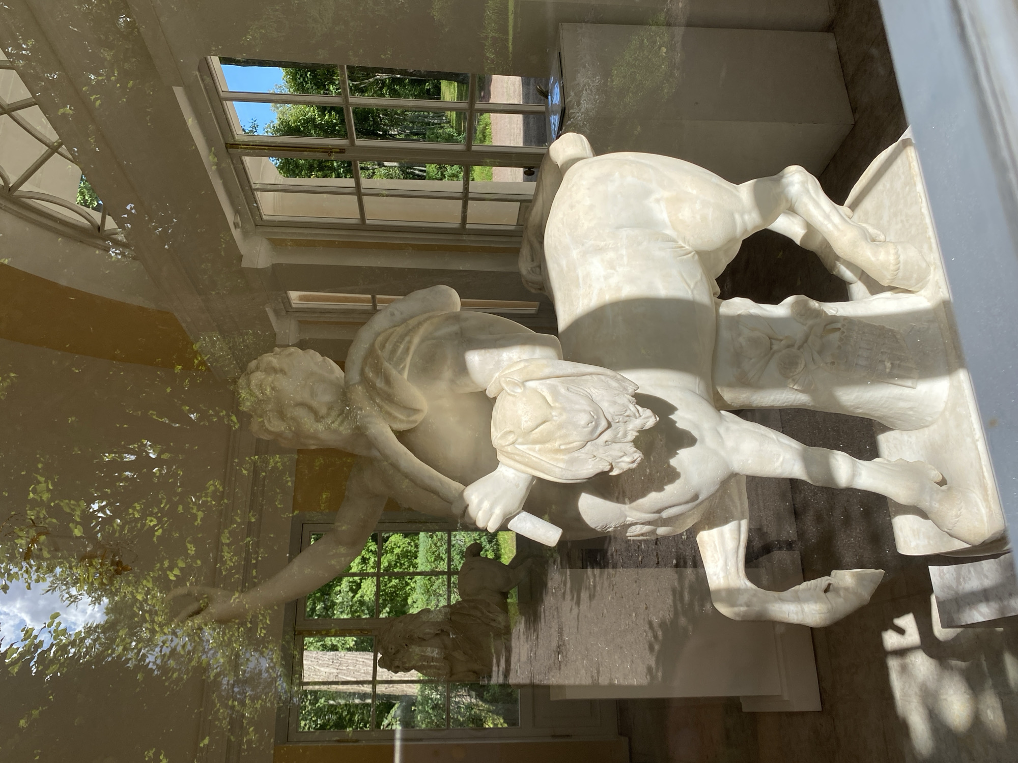 This is a photograph taken through a window, showing reflections on the glass surface, of a marble sculpture of a centaur inside a building. The centaur is depicted with the upper body of a human and the lower body of a horse. The sculpture is white and appears to be in good condition. It is standing on a pedestal. Behind the centaur, through the window, is a view of a lush green park with trees and a bright blue sky. Sunlight filters through the trees, casting shadows on the ground. There is another similar sculpture of a centaur visible in the background, further into the park. The building appears to be part of a palace or historical estate, with classical architectural elements visible in the interior, such as columns and large windows. The setting is likely the Pavlovsk Palace and Park ensemble, located in the city of Pavlovsk, Russia, a suburb of Saint Petersburg, known for its picturesque landscape park and imperial residences.