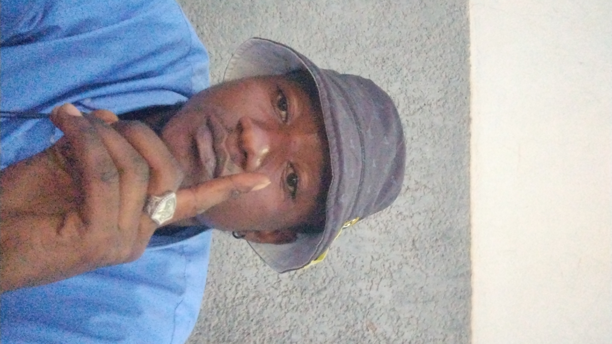 A person with dark skin, wearing a blue shirt and a dark-colored bucket hat, is shown indoors against a gray and white wall. The person is looking at the camera with their right index finger pointing toward their right eye. A ring is visible on the finger. The location is potentially within the city of Jos, Nigeria, though this cannot be definitively confirmed from the image.