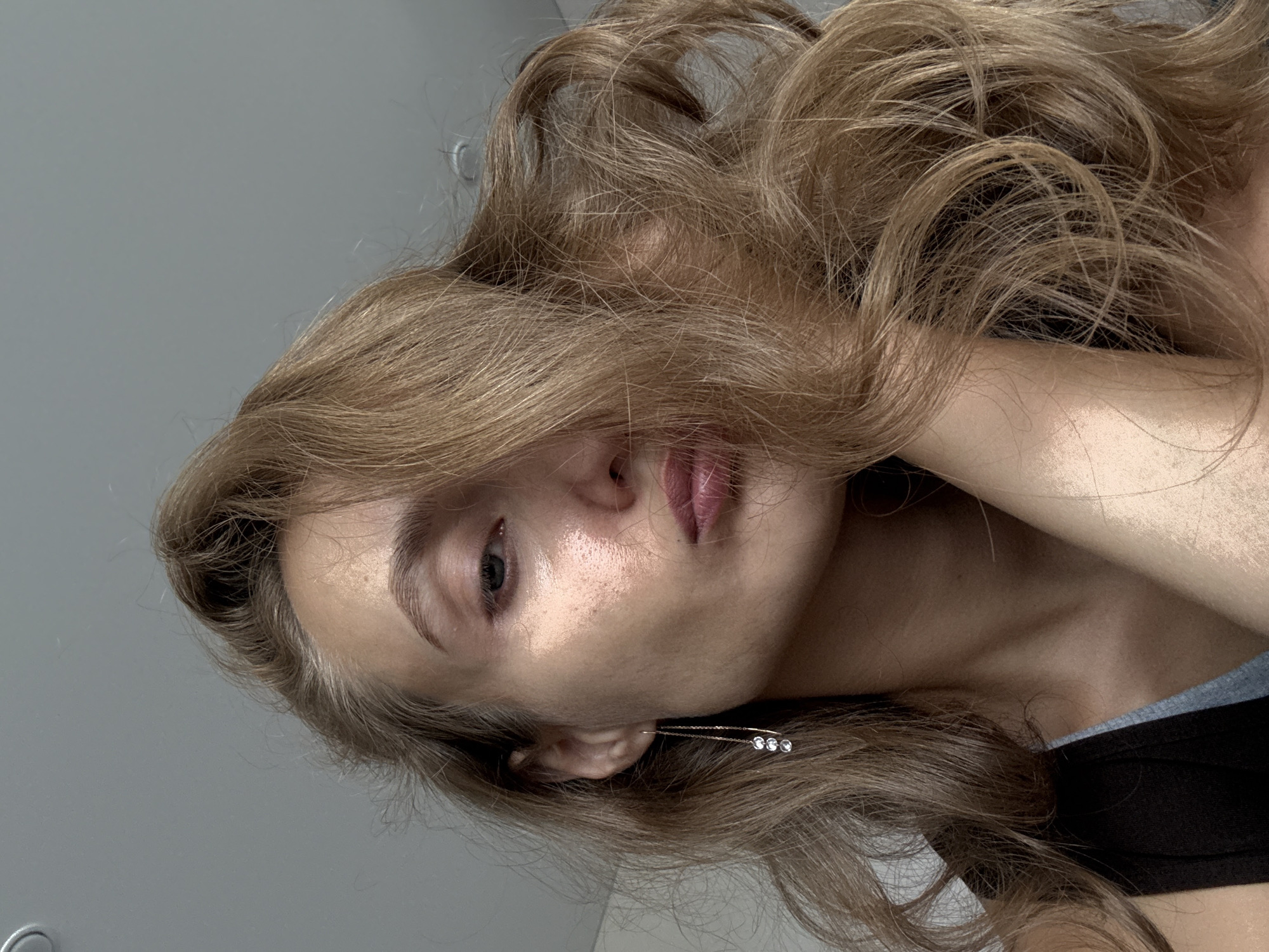 A close-up, low-angle portrait shows a woman's face from the nose up, with her right side more visible. Her blonde hair is tousled and partially obscures her face. She is wearing a dark tank top over a grey undershirt. A long earring with three clear stones dangles from her visible ear. Her skin appears to have a subtle sheen, possibly from makeup or sweat. Her lips are slightly pursed. The background is a plain, light gray wall, indicating an indoor setting. The image was taken in the city of Podshivalovo, Russia.