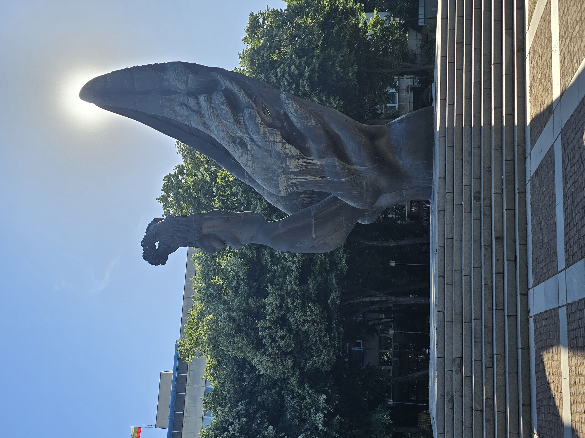A bronze statue of a bird in flight stands in Unknown, Unknown. The bird's head is obscuring the sun, resulting in a bright glare. The statue's wing and arm details are visible against a backdrop of green trees and a clear blue sky. To the right of the statue is a stone structure with vertical ridges and brickwork. The ground around the structure is paved. In the distance, a building is visible with a flag.