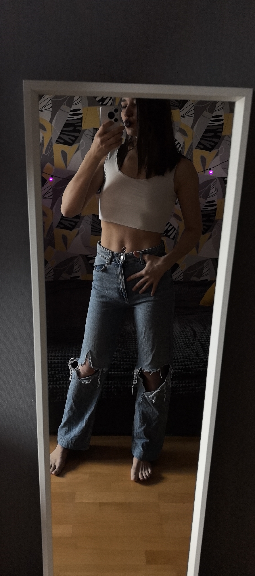 A full-body mirror selfie captures a person, likely a woman, holding a white smartphone to photograph her reflection. She has dark hair and is wearing dark lipstick. Her attire includes a white sleeveless crop top that exposes her midriff, where a belly button piercing is visible. She is also wearing light blue ripped jeans with prominent tears at both knees. The person is barefoot, standing on a light wooden floor.
In the reflection behind her, a wall covered in a distinctive wallpaper featuring abstract, leaf-like patterns in shades of grey, yellow, and black is visible. A dark-colored sofa or bed can be seen in the background reflection, and faint purple string lights are draped on the wall. One of her hands rests on her hip while the other holds the phone up to take the picture. The overall setting appears to be an indoor room, possibly a bedroom or living area. There is no visible text in the image.