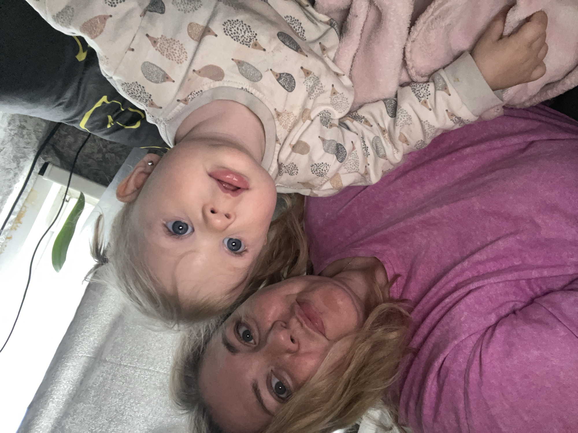 This close-up portrait, rotated 90 degrees clockwise, features an adult woman and a young child, likely a mother and her child, lying side-by-side in an indoor setting in Фоки, Russia. Both subjects are looking directly at the camera with soft, relaxed expressions.
The adult, positioned on the right side of the rotated image, has light blonde hair and blue-grey eyes, with a faint, gentle smile. She is wearing a textured, long-sleeved top in a vibrant magenta or purple hue.
The child, positioned to the left of the adult, has fair skin, light blonde hair, and striking blue eyes. They are wearing a cream-colored long-sleeved top patterned with numerous small brown and grey hedgehogs. A tiny silver stud earring is visible in the child's left earlobe. Their lips are slightly parted. A fluffy pink blanket or fabric is tucked close to the child's right arm and hand.
The background elements suggest a comfortable indoor environment. To the far left, a dark fabric, possibly a cushion or bedding, is visible. Below that, a light-colored, perhaps silver or grey, textured curtain or sheet adds to the soft surroundings. A green plant and some thin dark wires are also partially visible in the upper left portion of the frame. The overall atmosphere is intimate and serene. No text is visible in the image.