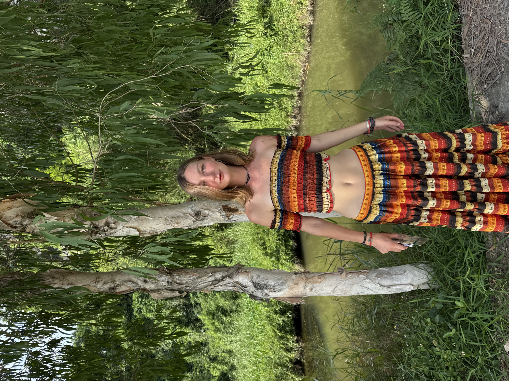 In the city of Nam Hoa Lư Ward, Vietnam, a young woman stands outdoors amidst lush greenery, posing for a photograph. She is positioned in front of a tranquil body of water, with trees and dense vegetation framing the scene. The woman is wearing a colorful, off-the-shoulder crop top and a matching long, tiered skirt. Her attire is adorned with a vibrant pattern of stripes and what appear to be animal motifs. She holds a small object, possibly a phone, in her right hand. The background reveals a natural landscape with a meandering waterway and abundant plant life, suggesting a serene, possibly rural, setting within the urban environment.