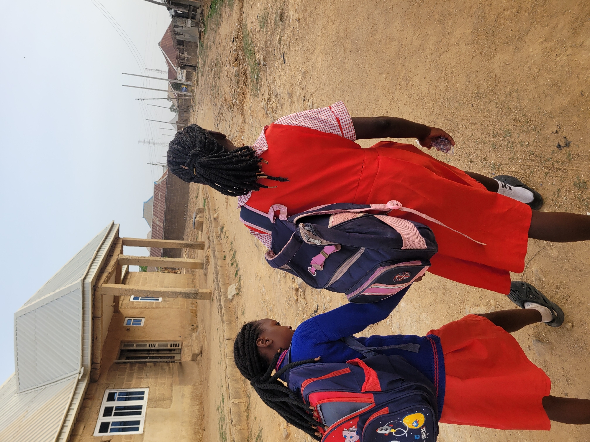 Two young girls in school uniforms are walking on a dirt road in Barakin Akawo, Nigeria. The girls are wearing red skirts and tops, with the younger girl in a blue sweater. They both have backpacks on. The surrounding environment includes a building, power lines, and other structures, indicative of a developing urban area. The ground is dusty and barren.