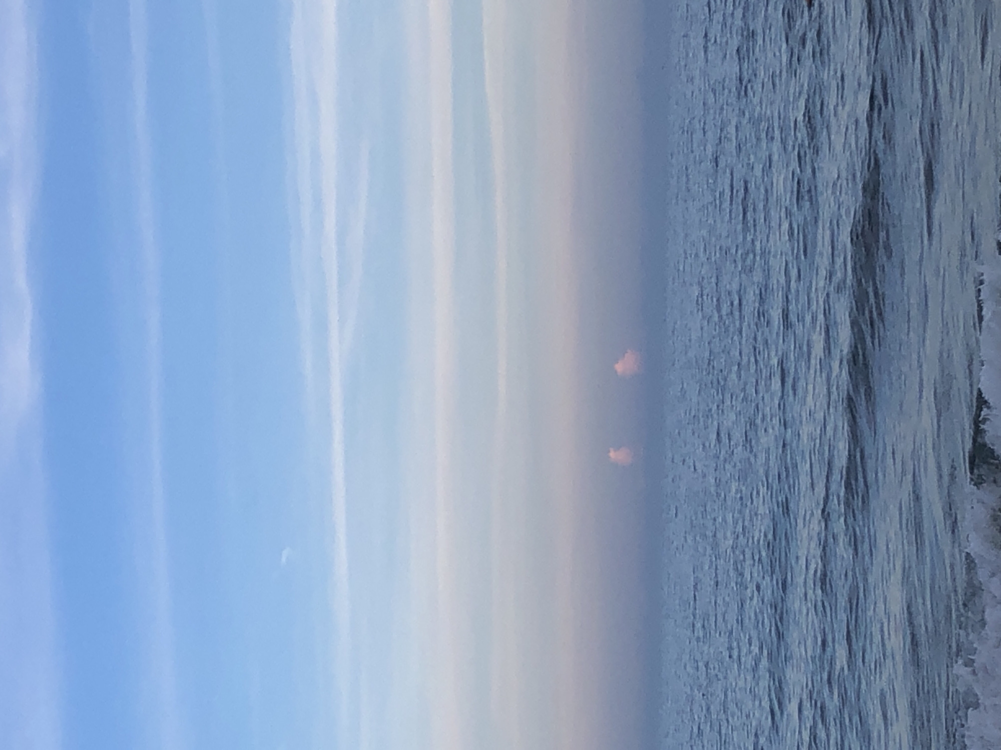 The image depicts a serene seascape with a clear horizon. The sky transitions from a light blue at the top to softer pastel hues of pink and lavender closer to the water. Two small, puffy clouds, tinged with pink, are visible on the horizon. The ocean below is a deep blue with gentle ripples and a foamy wave breaking in the foreground. The overall impression is one of tranquility. There are no people or man-made objects clearly visible in the scene, suggesting a natural, undeveloped coastal environment. The lack of specific landmarks makes it impossible to definitively identify the location as Phu Thuy Ward, Vietnam based solely on the visual information provided.