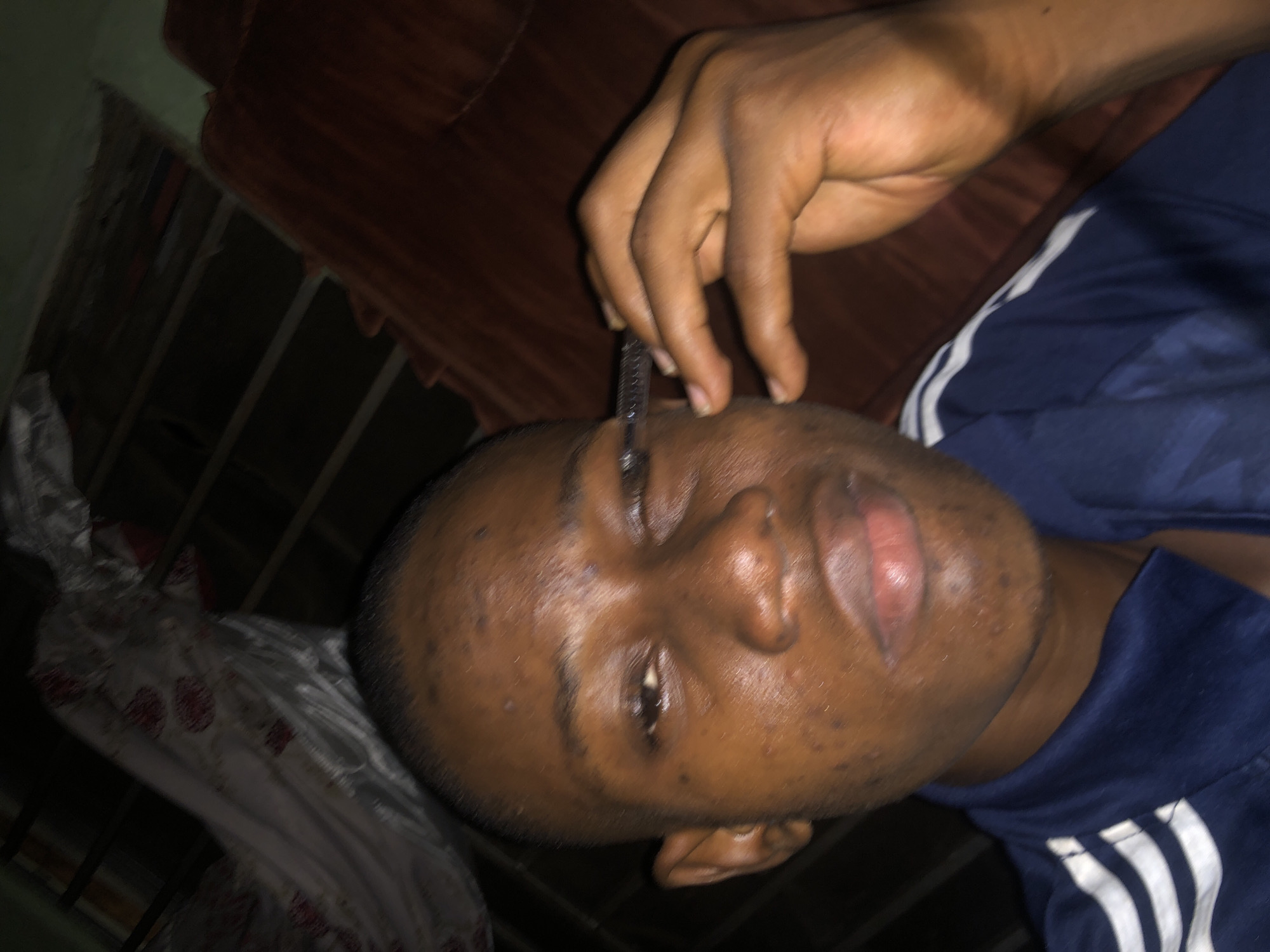 A person lies on their back in an indoor setting. The person is dark-skinned, wearing a blue shirt with white stripes on the sleeves. A hand holds a black object near the person's face, possibly touching the forehead. The background shows a brown object and a white, patterned object. The setting is indoors. The location in the city of Atani, Nigeria is unknown.