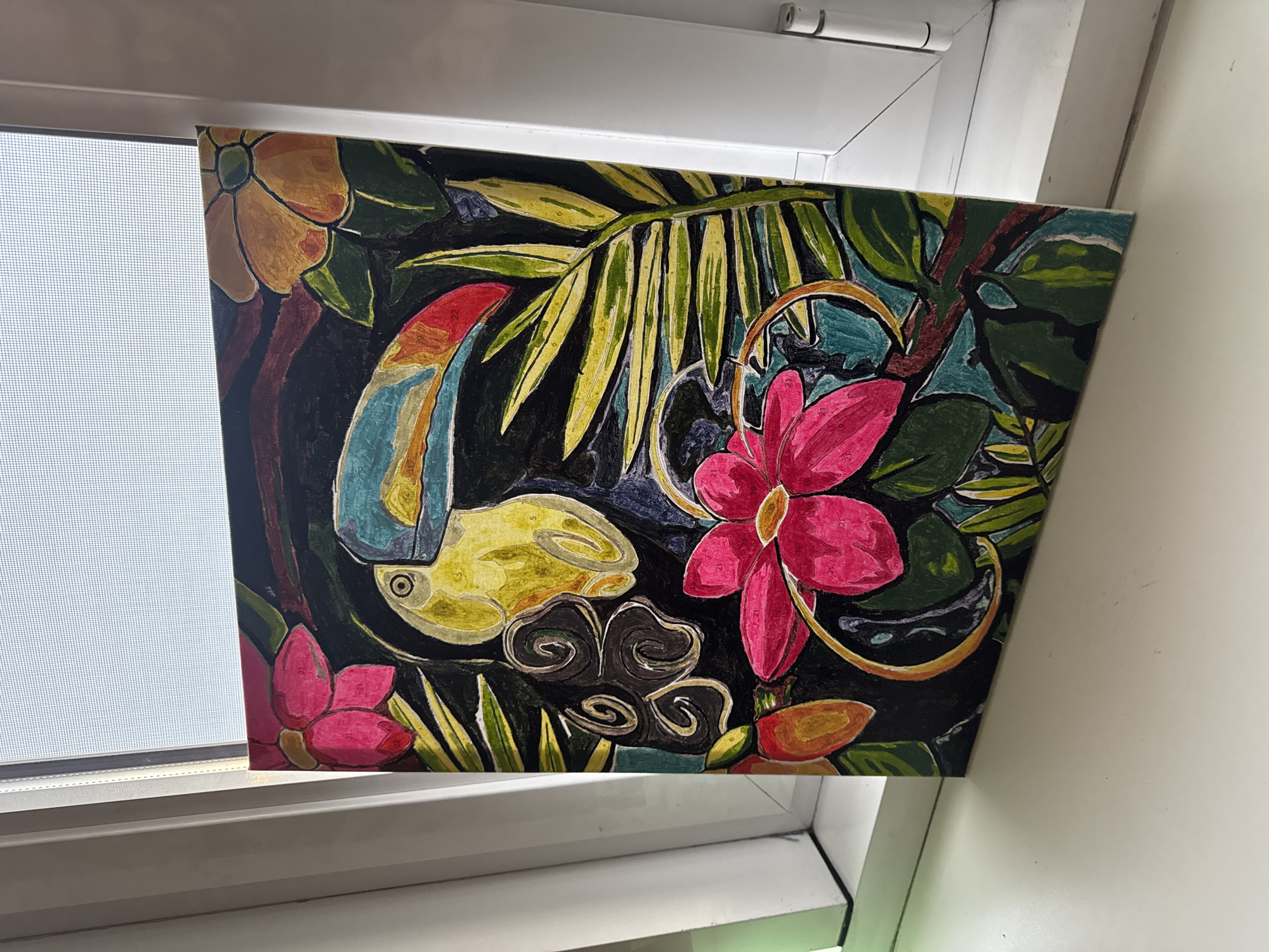 The image shows a painting of a toucan surrounded by vibrant flowers and foliage, placed on a white windowsill. The painting is colorful, with a black background that makes the toucan and the surrounding elements stand out. The toucan is yellow with a large, multicolored beak. There are pink and orange flowers, as well as green leaves and palm fronds. The painting is angled slightly, leaning against the window. The window itself has a screen visible through it, suggesting an indoor setting. The windowsill is clean and white, and the overall scene is well-lit by natural light coming from the window. There are no people or animals present in the image, and no indication of the city of Almaty, Kazakhstan.