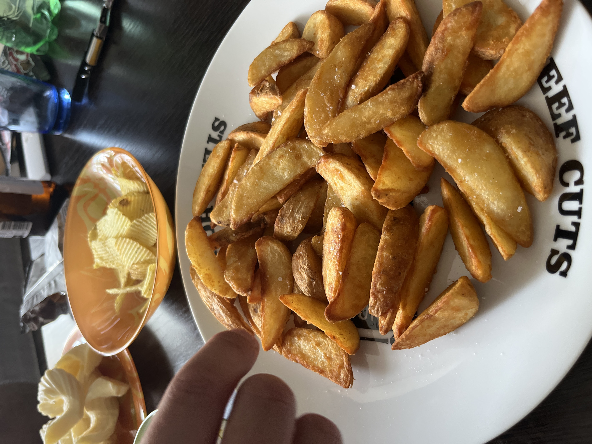 This image, taken in Rusanovo, Russia, depicts a close-up view of a dining table laden with food and beverages. In the immediate foreground, a white, round plate is filled with numerous golden-brown potato wedges, visibly seasoned with coarse salt crystals. The plate features black, stylized text, partially visible and oriented upside down, spelling "BEEF CUTS." A human hand, showing two fingers, extends from the upper-left towards the potato wedges, positioned as if about to grasp one.
To the left, slightly out of focus, is a smaller, patterned plate holding several light yellow, elaborately shaped items, consistent with butter curls or similar decorative food elements. Adjacent to this, in the midground, an orange, translucent bowl contains a quantity of crinkle-cut potato chips.
The background reveals a dark, presumably wooden, table surface. Various objects are arranged on it: a brown glass bottle, partially visible; a clear blue drinking glass; a green plastic bottle; and a slender, black and metallic object, resembling a vape pen or a ballpoint pen. The overall setting appears to be an indoor dining environment, possibly a kitchen or dining room.