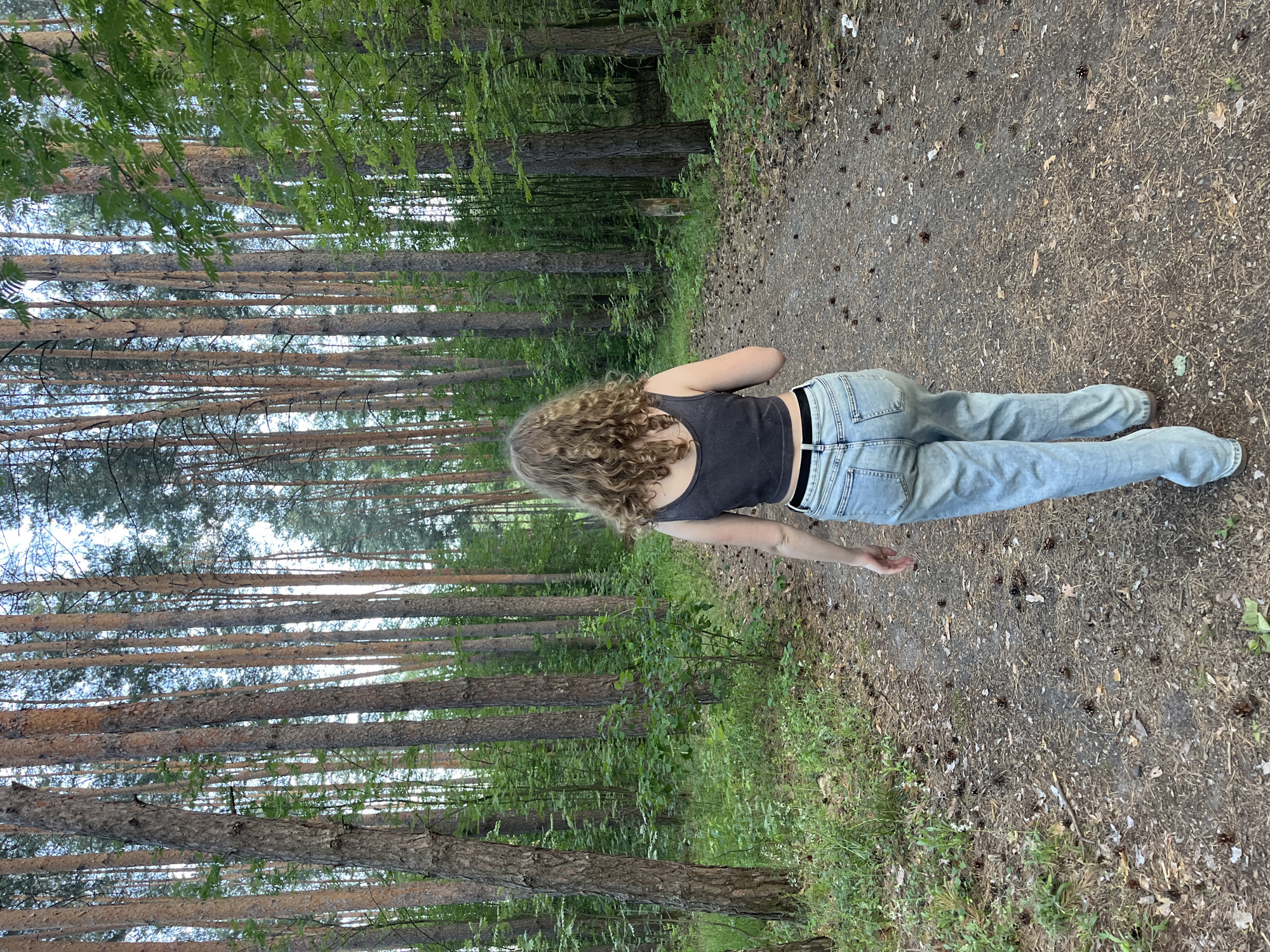 A young woman with curly blonde hair walks away from the camera down a dirt path in a pine forest. She is wearing a dark gray tank top and light blue jeans. The path is covered in pine needles and pinecones, with tall pine trees on either side. Sunlight filters through the canopy, illuminating parts of the forest floor. The scene is located in a wooded area, likely a park or nature reserve.