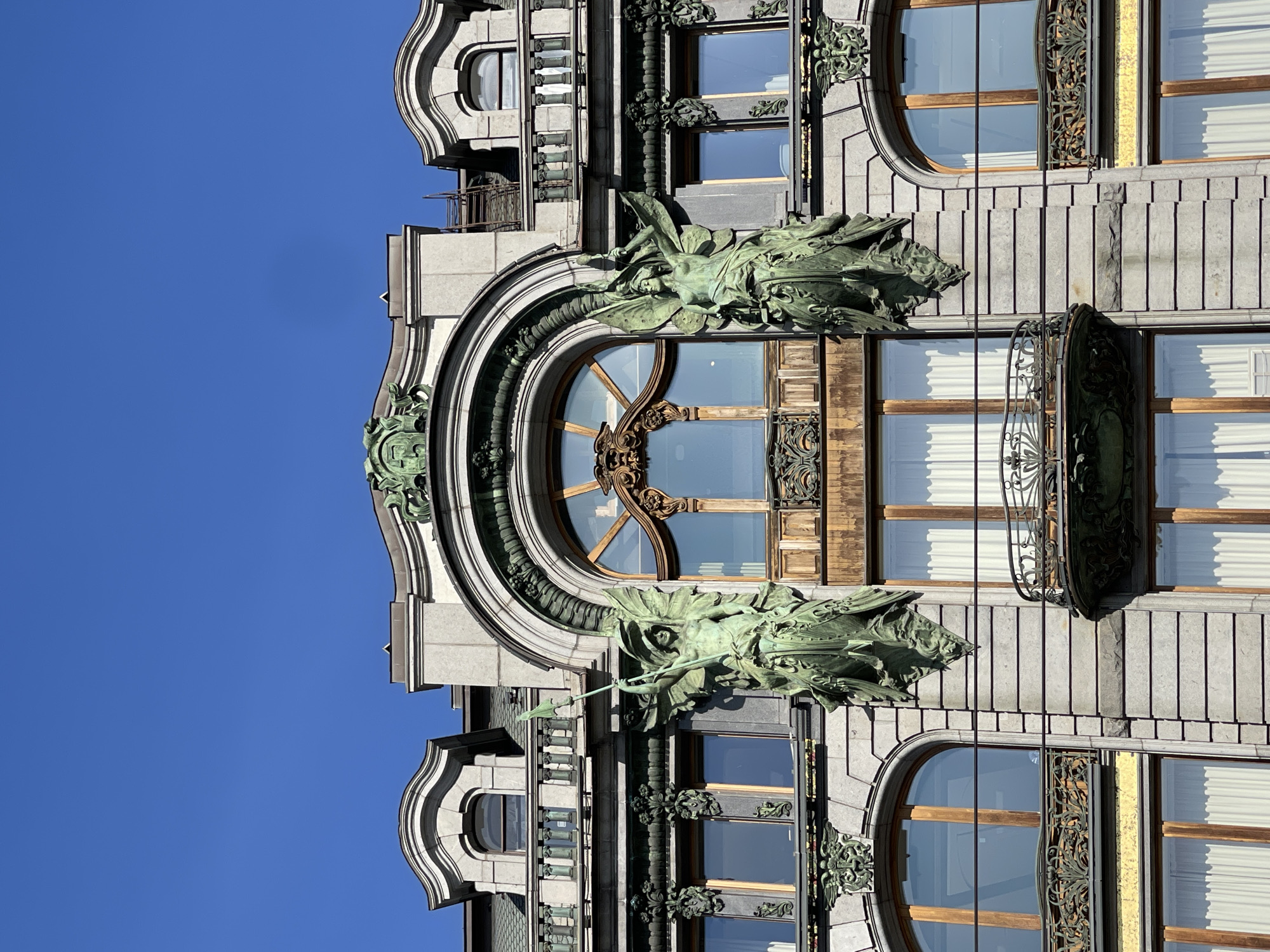 The image depicts a section of the Singer House (Dom Zinger), an Art Nouveau building located on Nevsky Prospekt in Saint Petersburg, Russia. The facade is adorned with architectural elements, including an arched window with intricate woodwork. On either side of the window, there are sculptural figures of women, seemingly made of bronze or a similar patinated metal. The building's facade is grey and features decorative details, including carved floral patterns and wrought-iron balconies. The sky is clear and blue, providing a backdrop for the building. The image does not feature people or actions.