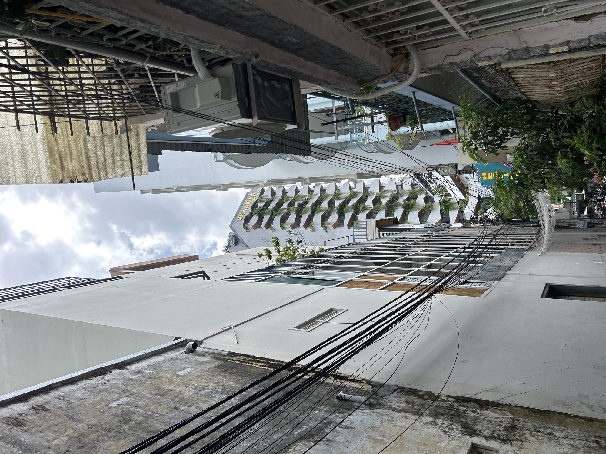 The image, captured from a low angle looking upwards, depicts a dense urban street or alleyway in Nha Trang, Vietnam, beneath an overcast daytime sky. The scene is dominated by multiple-story buildings and a complex web of overhead cables.
In the immediate foreground and to the left, an older building with a light-colored concrete facade shows signs of weathering. Numerous thick, black electrical or communication cables crisscross the frame, trailing down this building's side and spanning the narrow gap between structures, creating a prominent and intricate visual element.
Dominating the mid-ground is a modern, multi-story building distinguished by a facade featuring staggered, horizontal planter boxes, each containing green foliage. The word "SEASIDE" is clearly visible in large, light-colored letters on its exterior, with additional, partially obscured text below it, possibly "MAX." Further down the street on the right, a partial green sign with yellow lettering, likely "HEL..." suggests a business, possibly a hotel.
Other visible objects include metal grating structures, possibly for shade or security, and external air conditioning units mounted on building walls. Patches of natural greenery, including small trees and potted plants, are seen on balconies and at street level on the right, where subtle splashing water indicates a fountain or water feature. The sky is predominantly cloudy, suggesting an overcast day with no direct sunlight. No people are visible, and no specific activities are taking place in this static view of the city's architecture.