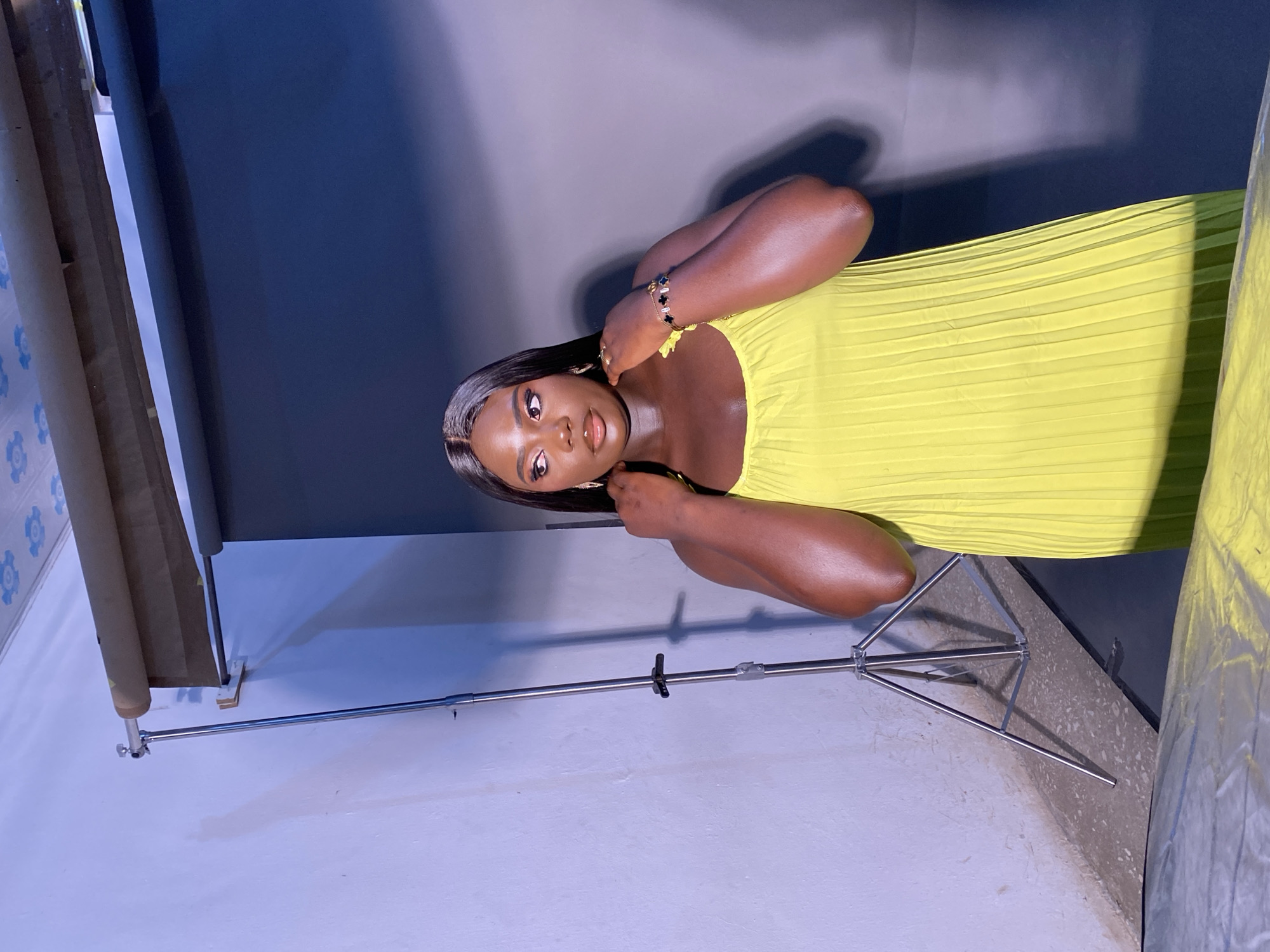 An overhead shot captures a young Black woman standing in what appears to be a studio. She is wearing a vibrant, pleated yellow dress and is posed with her hands touching her neck, looking away from the camera. A silver tripod stands to her left, and a dark gray backdrop is behind her, contrasted by a light-colored wall on the left side of the frame. The scene is lit by studio lights, casting shadows on the wall and floor. The woman has long, dark hair and is adorned with jewelry, including earrings and a bracelet.
