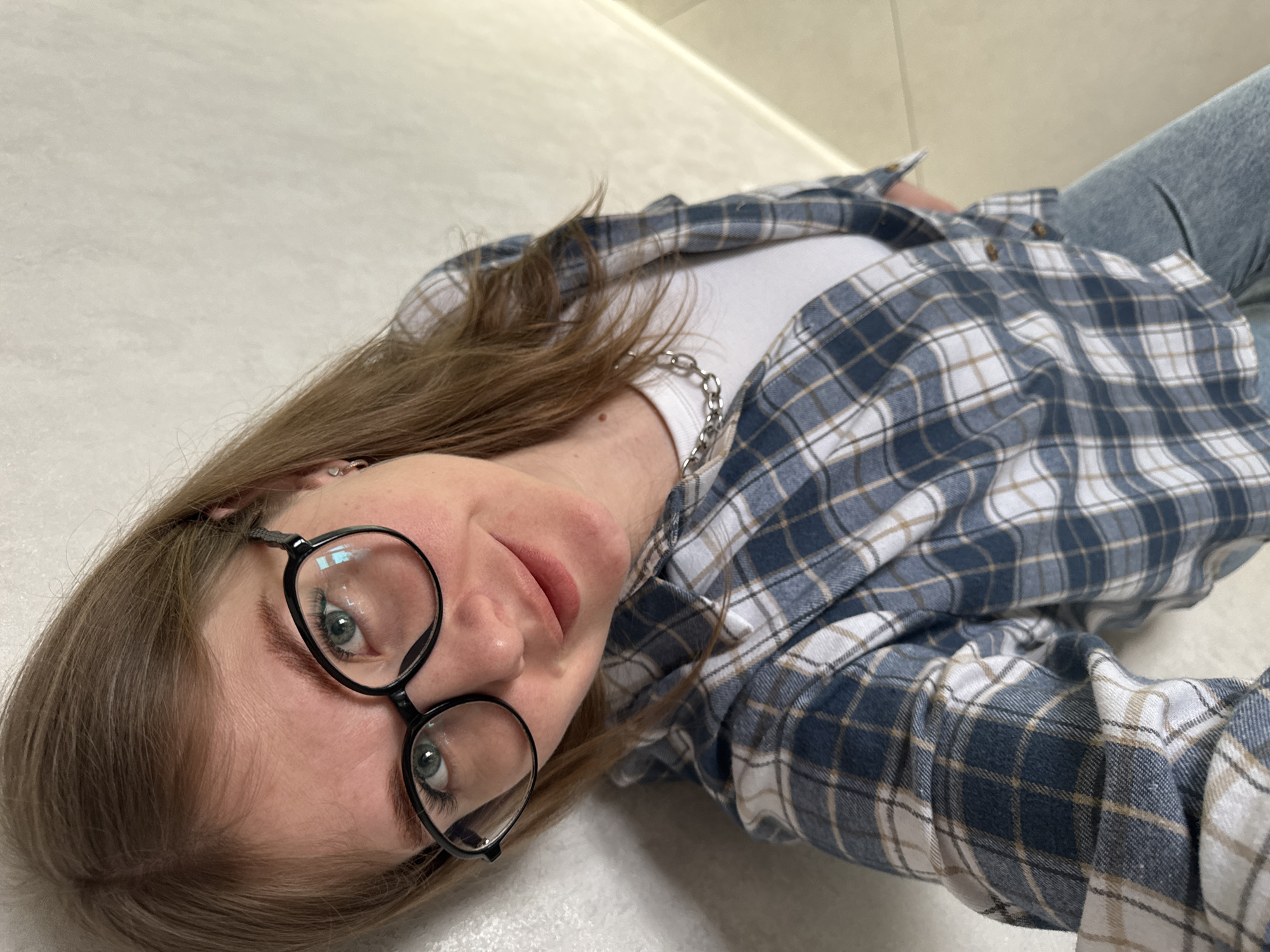 A female individual is depicted in a high-angle, close-up indoor portrait in Gornyi (Dvurechenskoe S/P), Russia. She is leaning against a textured wall, which transitions from off-white on the left to a light beige on the right. The subject has light brown hair, wears black-framed eyeglasses, and displays light green/hazel eyes with a faint smile. She is dressed in a white t-shirt, an unbuttoned blue, white, and tan plaid shirt, and light blue denim jeans. A silver-colored chain necklace is visible over the t-shirt, and a small silver hoop earring is present in her left ear. Her gaze is directed towards the camera.
